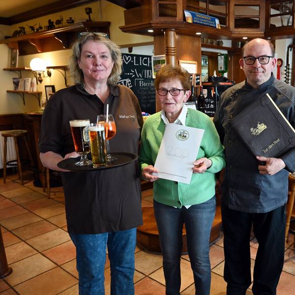 Ein Mann, eine Frau und die Mutter des Mannes stehen gemeinsam in der Gaststube und vor der Theke eines Hotel-Restaurants.