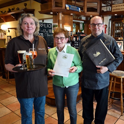 Ein Mann, eine Frau und die Mutter des Mannes stehen gemeinsam in der Gaststube und vor der Theke eines Hotel-Restaurants.