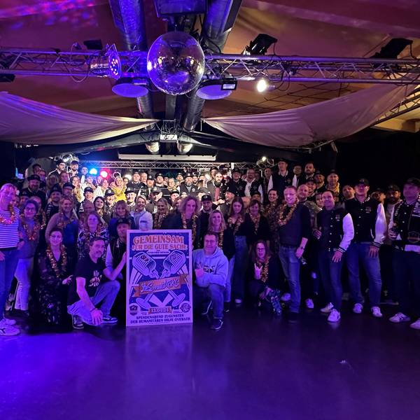 Zwei Chöre stehen im blauen Licht im Saal Altenrath in Marialinden und halten ein Plakat für ein Benefizkonzert.
