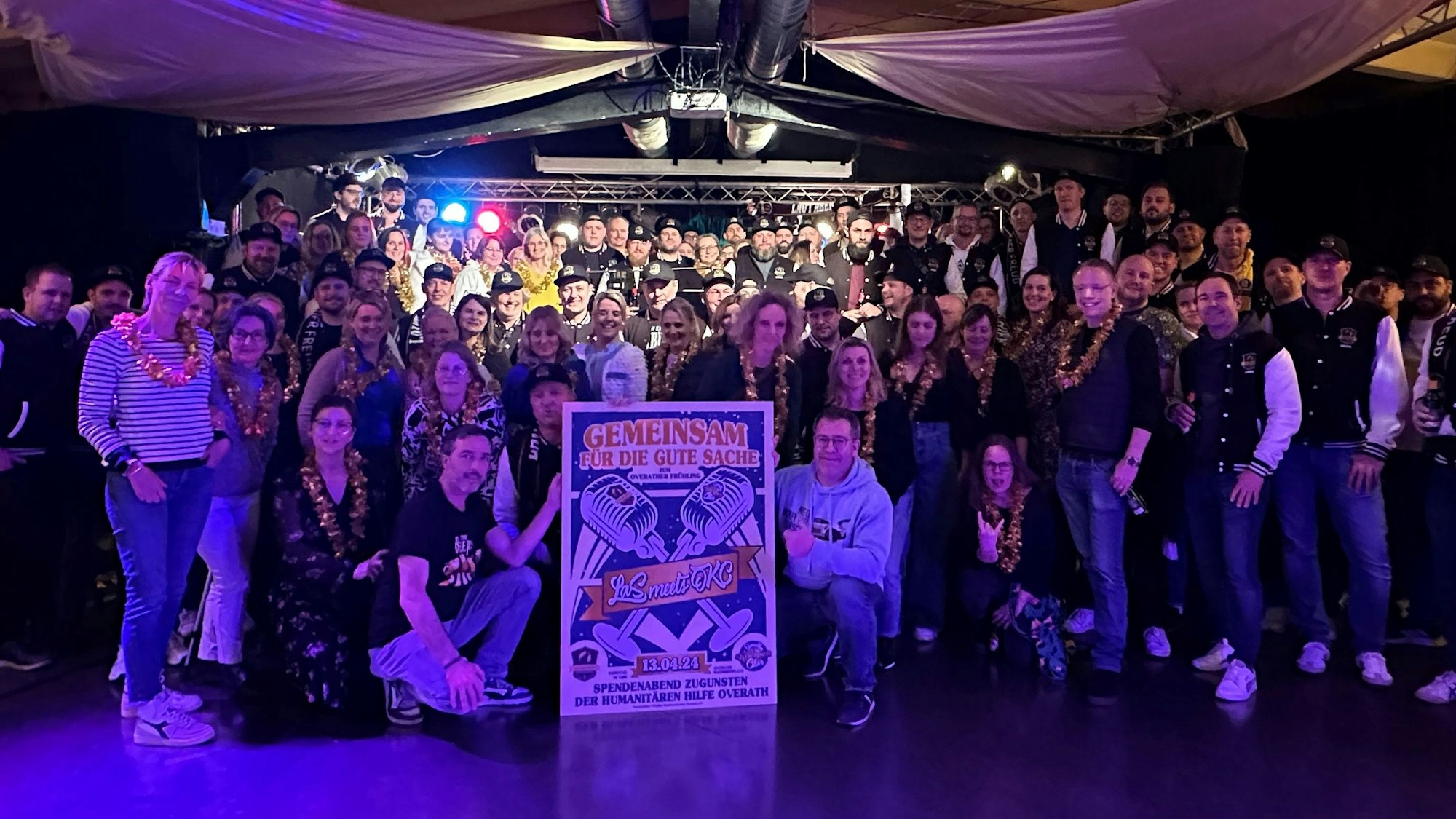 Zwei Chöre stehen im blauen Licht im Saal Altenrath in Marialinden und halten ein Plakat für ein Benefizkonzert.