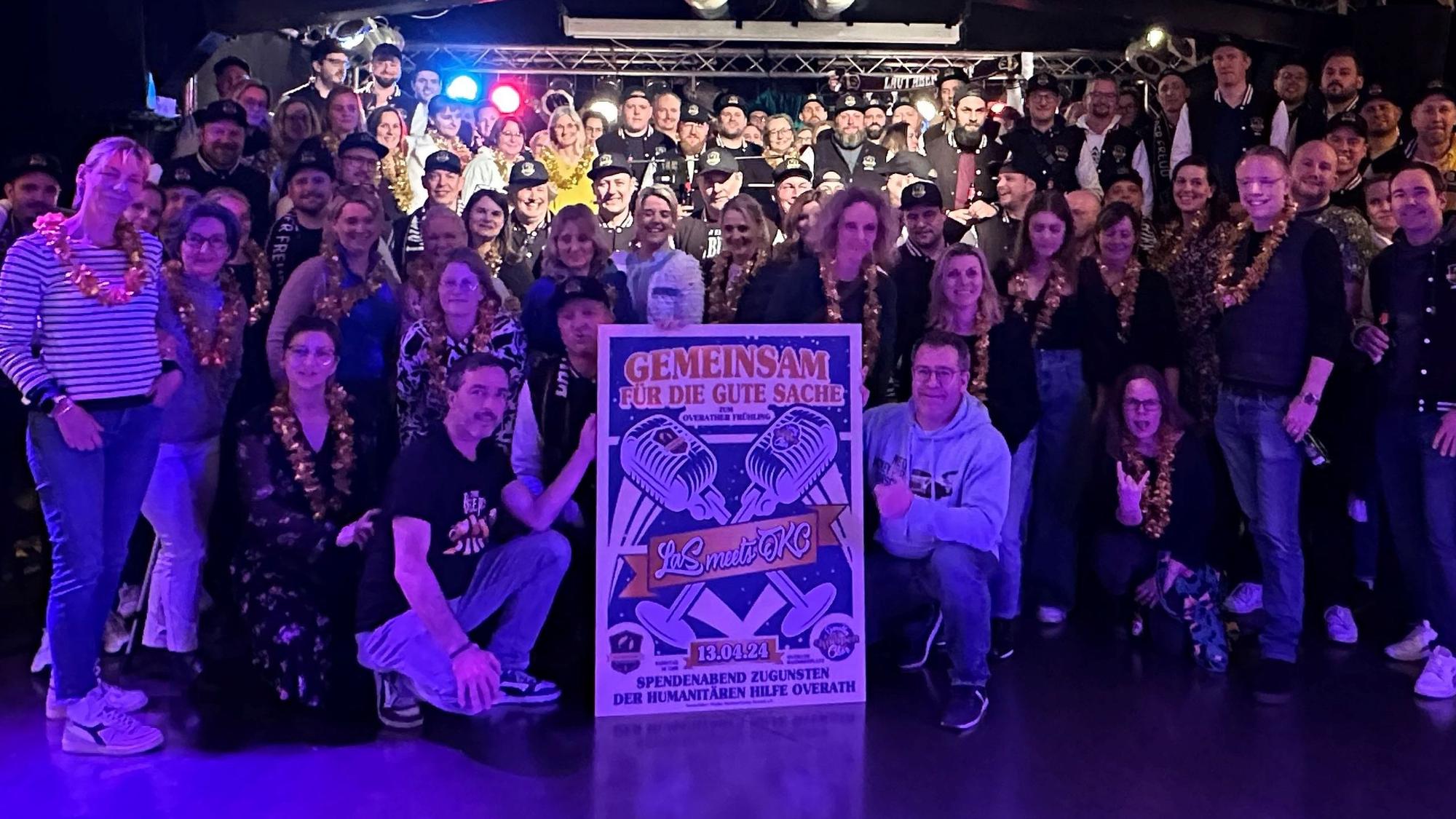 Zwei Chöre stehen im blauen Licht im Saal Altenrath in Marialinden und halten ein Plakat für ein Benefizkonzert.