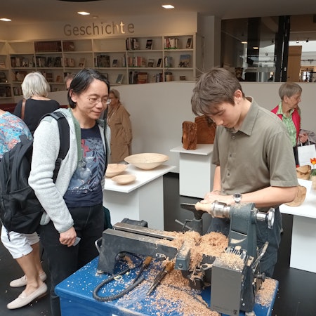 Ein junger Mann steht an einer Holzbearbeitungsmaschine, zwei Frauen gucken ihm zu.