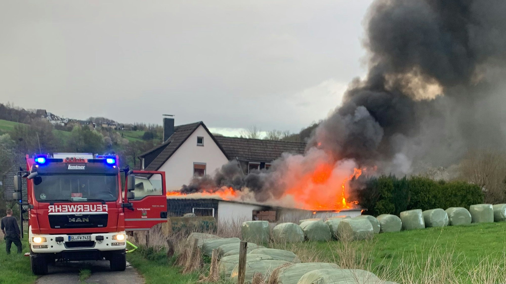 Ein Haus brennt in voller Ausdehnung, daneben steht ein Feuerwehrfahrzeug mit Blaulicht auf einer schmalen Straße.