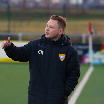 Trainer Christopher Kockerols steht an der Seitenlinie eines Fußballplatzes und hält den Daumen der rechten Hand nach oben.