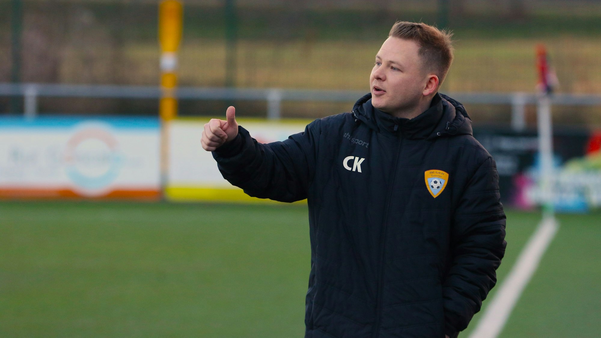Trainer Christopher Kockerols steht an der Seitenlinie eines Fußballplatzes und hält den Daumen der rechten Hand nach oben.
