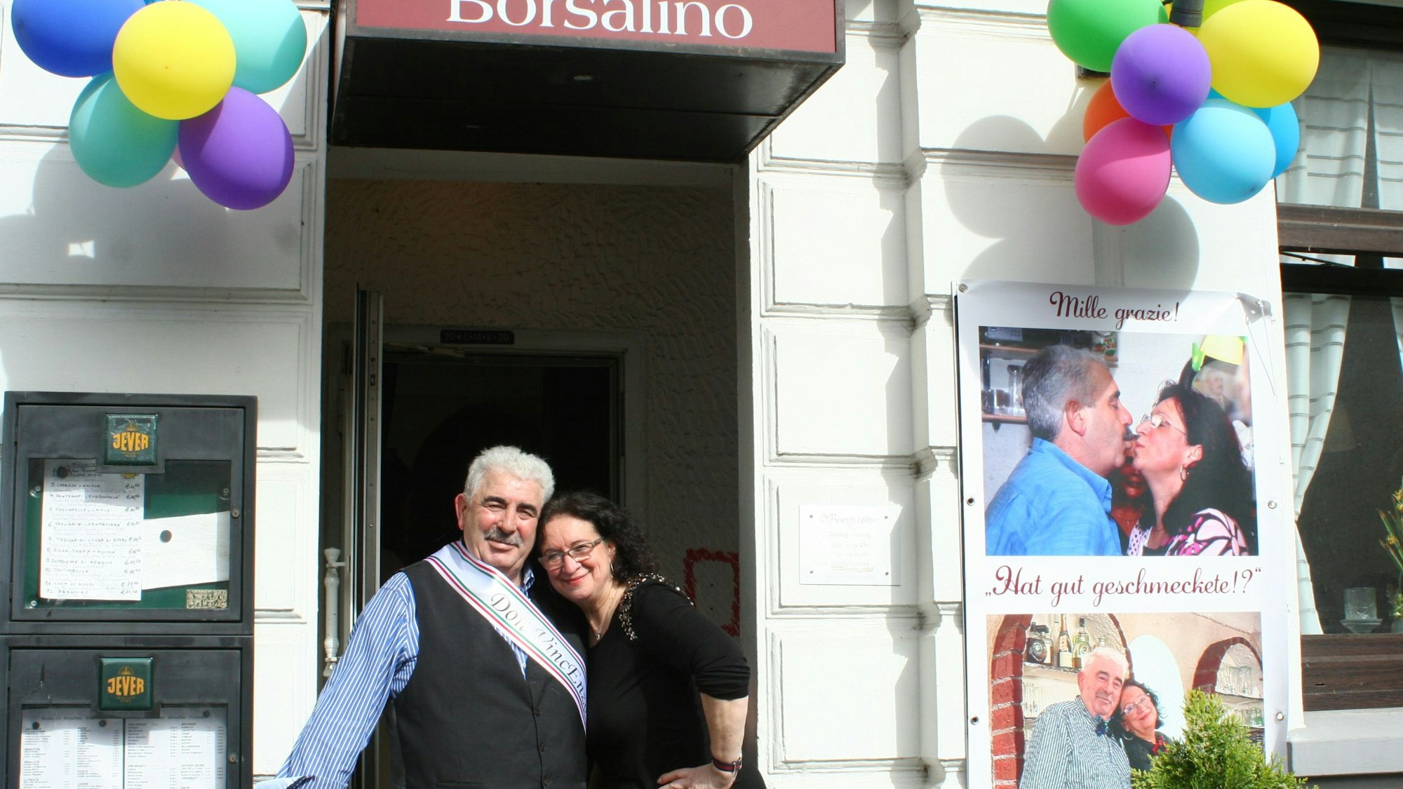 Ein grauhaariger Mann und eine dunkelhaarige Frau stehen in einem Hauseingang, er trägt eine Schärpe.