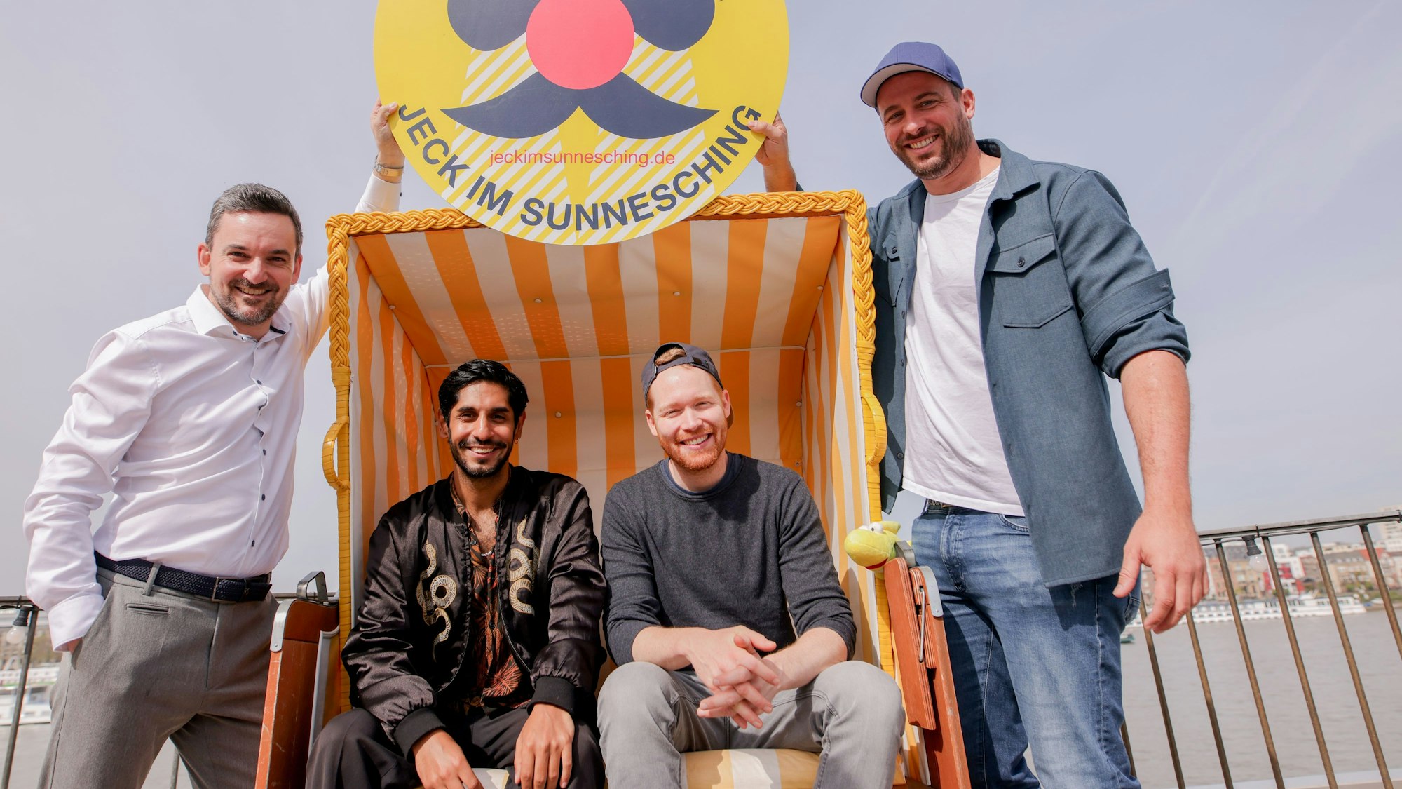 Björn Lindert, geschäftsführender Gesellschafter x4kids, Juri Rother (Planschemalöör), Nils Schreiber (Miljö) und Martin Becker, Geschäftsführer Sharkys Schwimmschule sitzen am Rhein in einem Strandkorb.