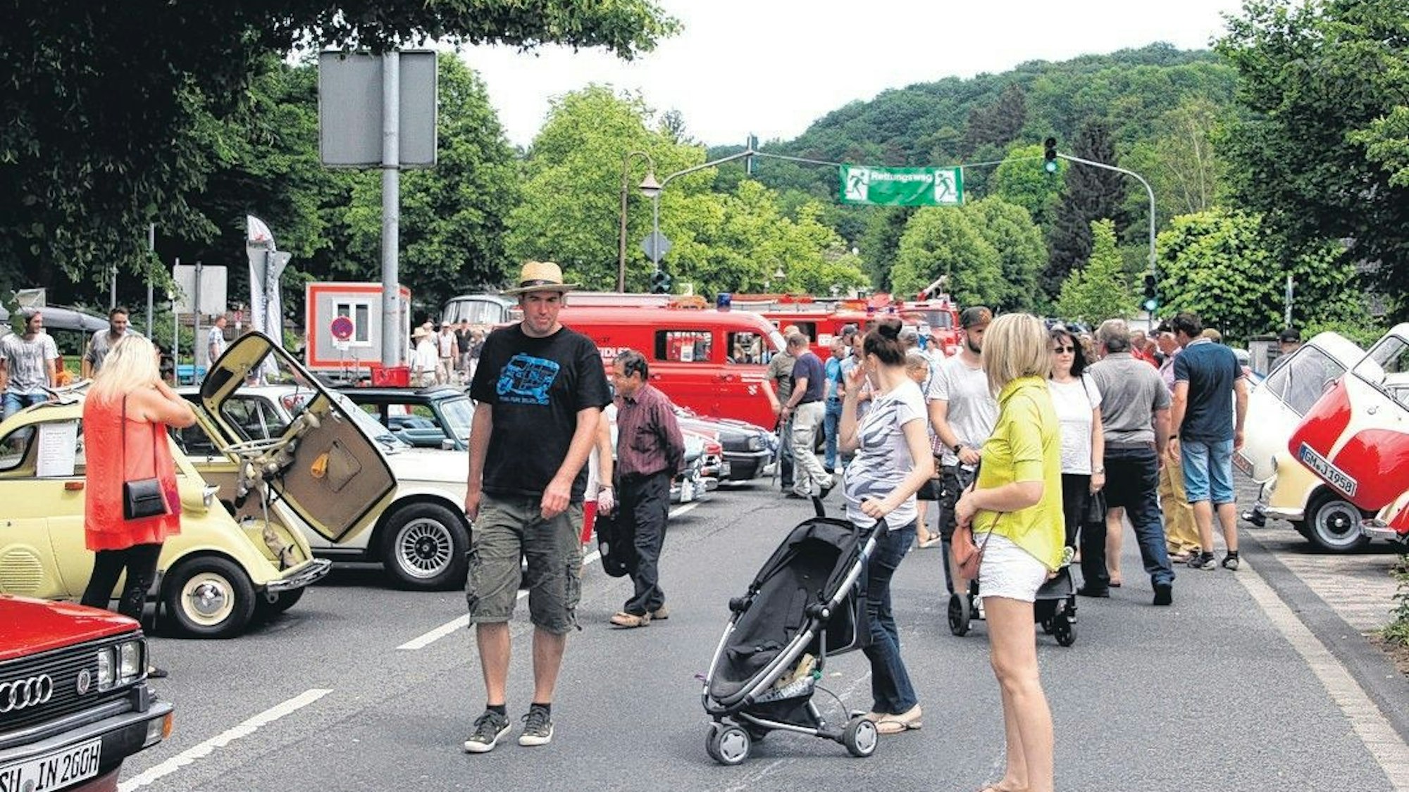Menschen in sommerlicher Kleidung schauen sich die in einer Reihe aufgestellten Oldtimer an.