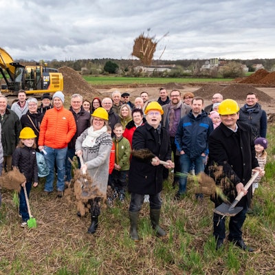 Ehrengäste des Baubeginns werfen beim ersten Spatenstich Erde in die Luft, dahinter weitere Vertreter von Politik, Verwaltung und Schulgemeinde. Auf der künftigen Baustelle steht auch schon ein Bagger.