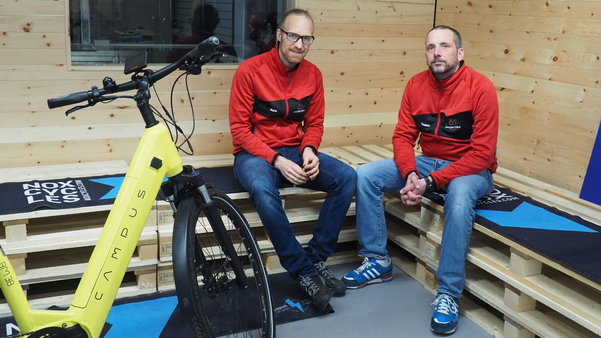 Zwei Männer sitzen auf Bänken, die aus Holz-Paletten gebaut sind. Vor ihnen steht ein gelbes Fahrrad.