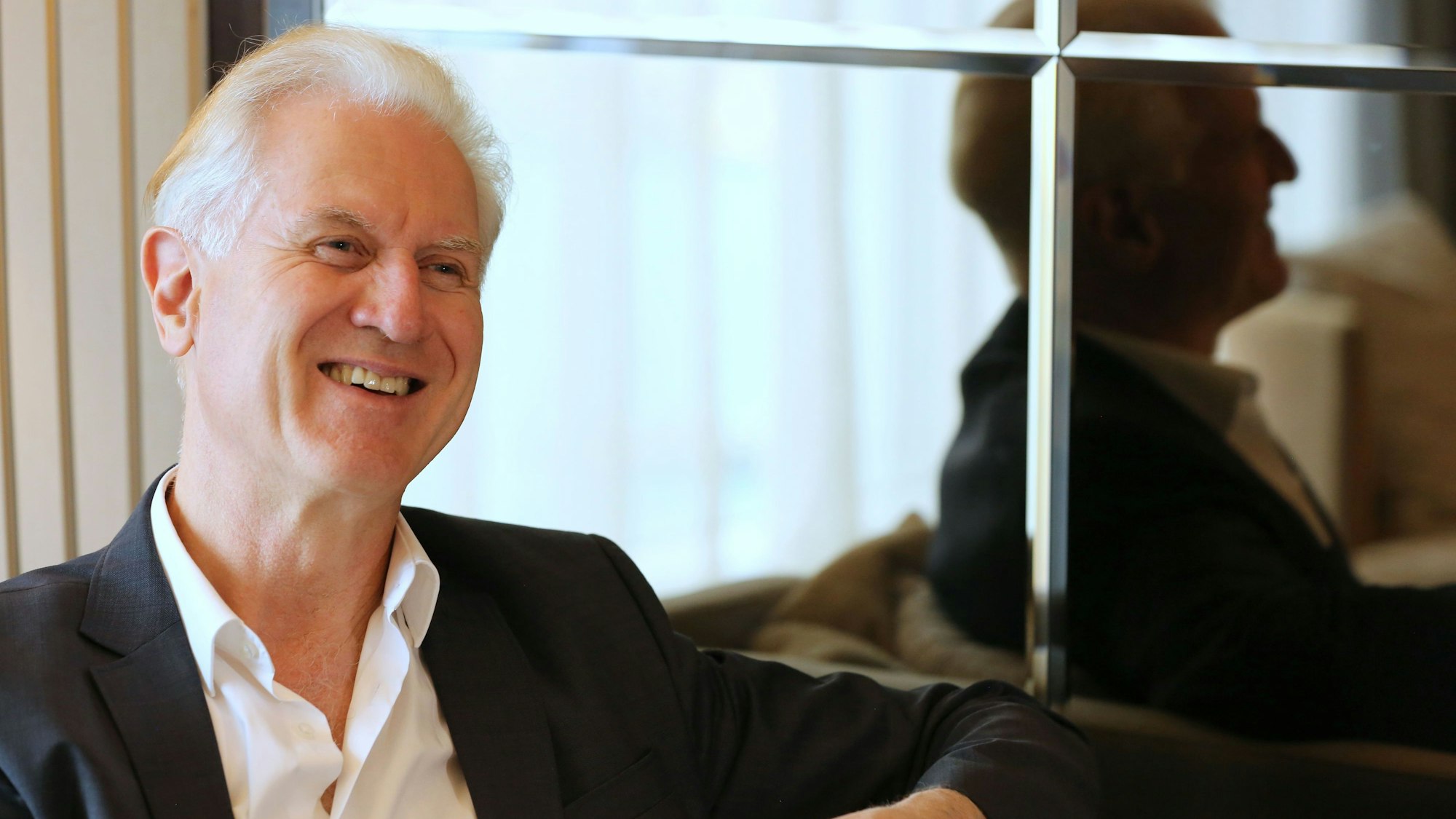 Chefdirigent Christoph Poppen sitzt auf einem Sessel und blickt freundlich lächelnd. Er trägt ein weißes Hemd und ein schwarzes Sakko.