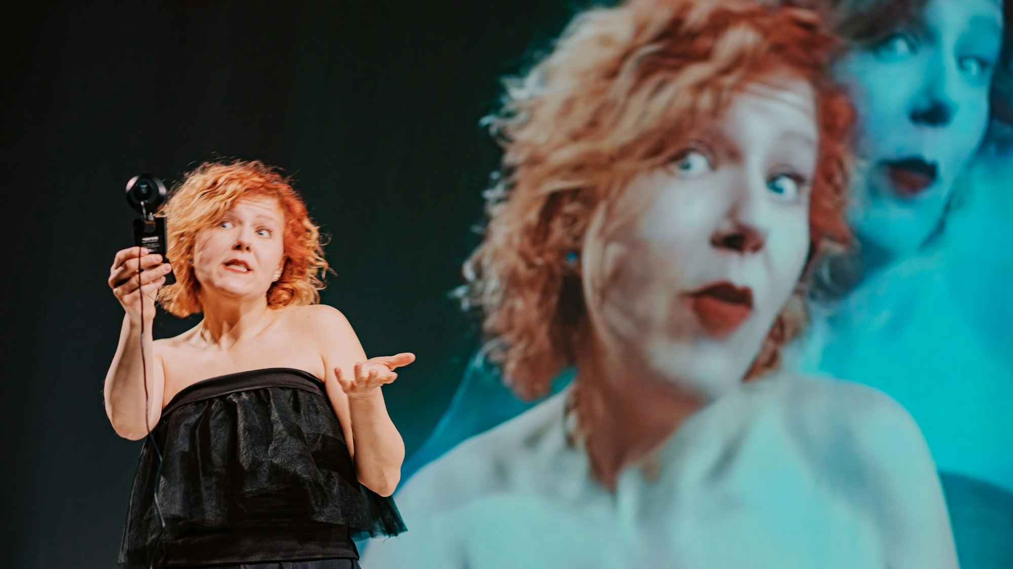 Frauke Albert hat rote Haare und trägt ein schwarzes Kleid. Hinter ihr werden Nahaufnahmen von ihr auf eine Leinwand projiziert, die aber zunehmend einen Blaustich bekommen.