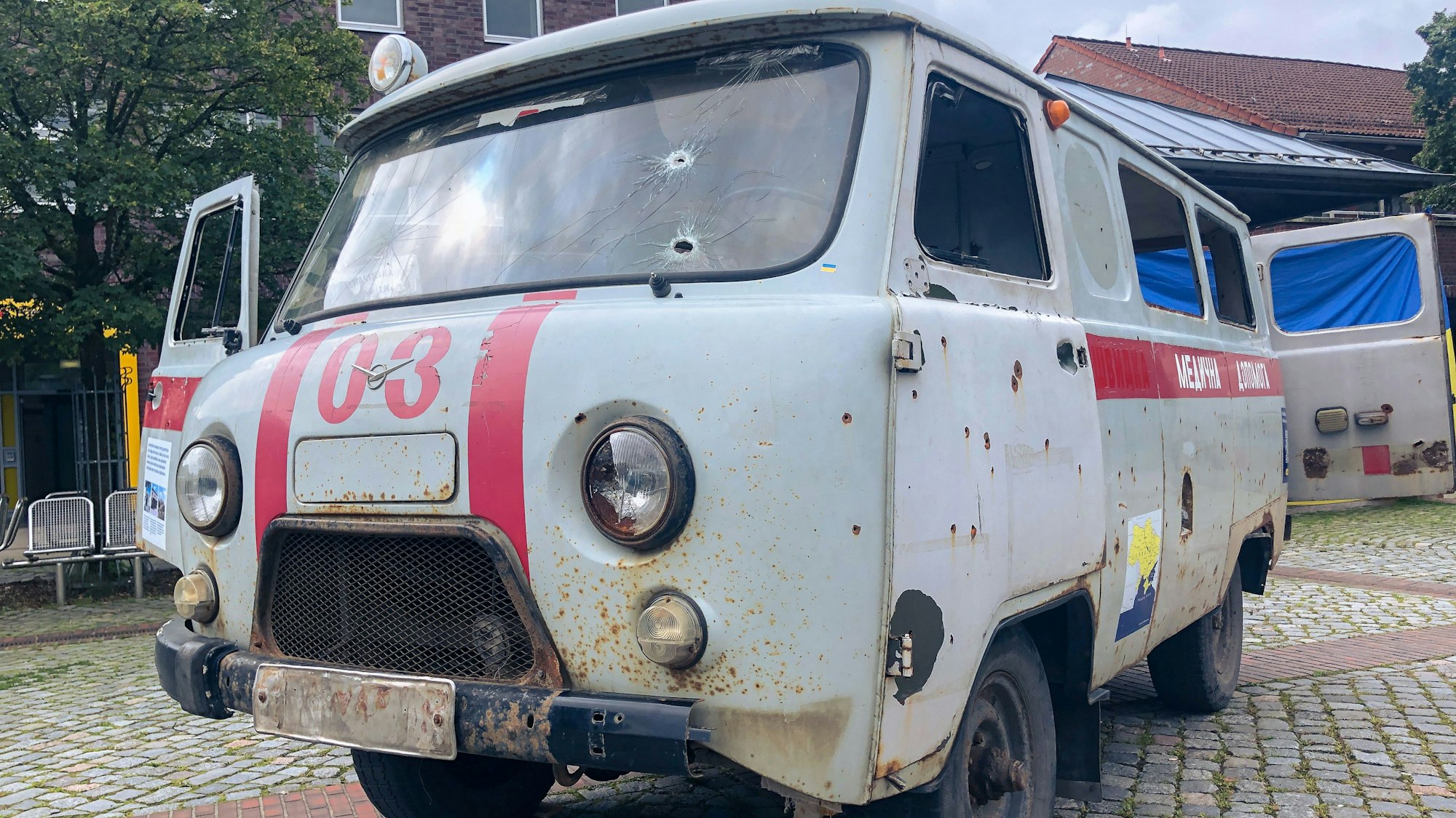 Ein Krankenwagen mit Einschusslöchern steht auf einem Platz.