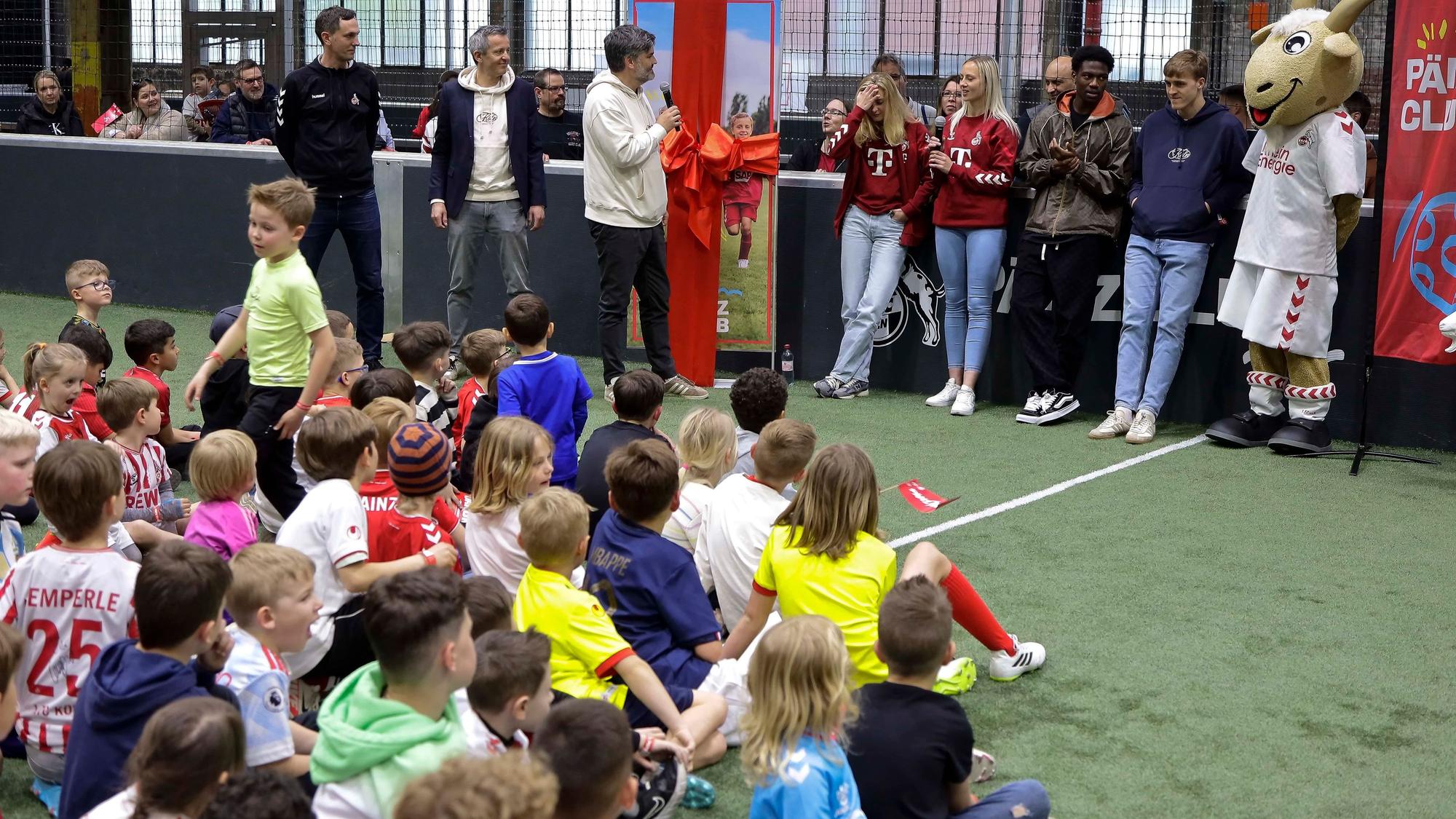 Zu sehen sind eine Gruppe von Kindern, die auf einem Fußballplatz sitzt und eine Gruppe von Erwachsenen, die an einer Wand steht.