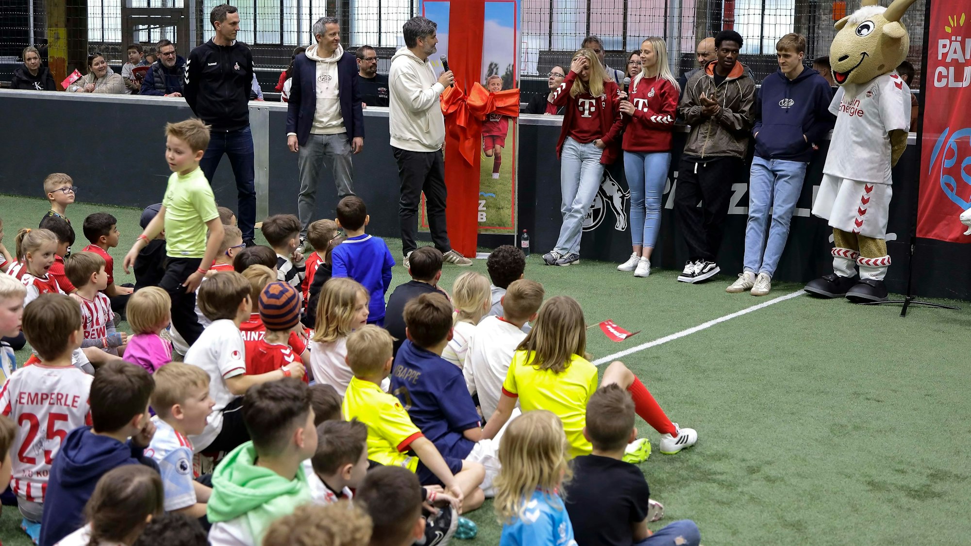 Zu sehen sind eine Gruppe von Kindern, die auf einem Fußballplatz sitzt und eine Gruppe von Erwachsenen, die an einer Wand steht.