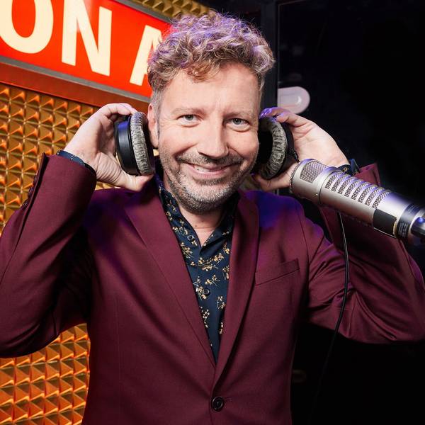 Thorsten Schorn steht an einem Mikrofon, er hat Kopfhörer in der Hand und trägt einen bordeauxfarbenen Anzug.