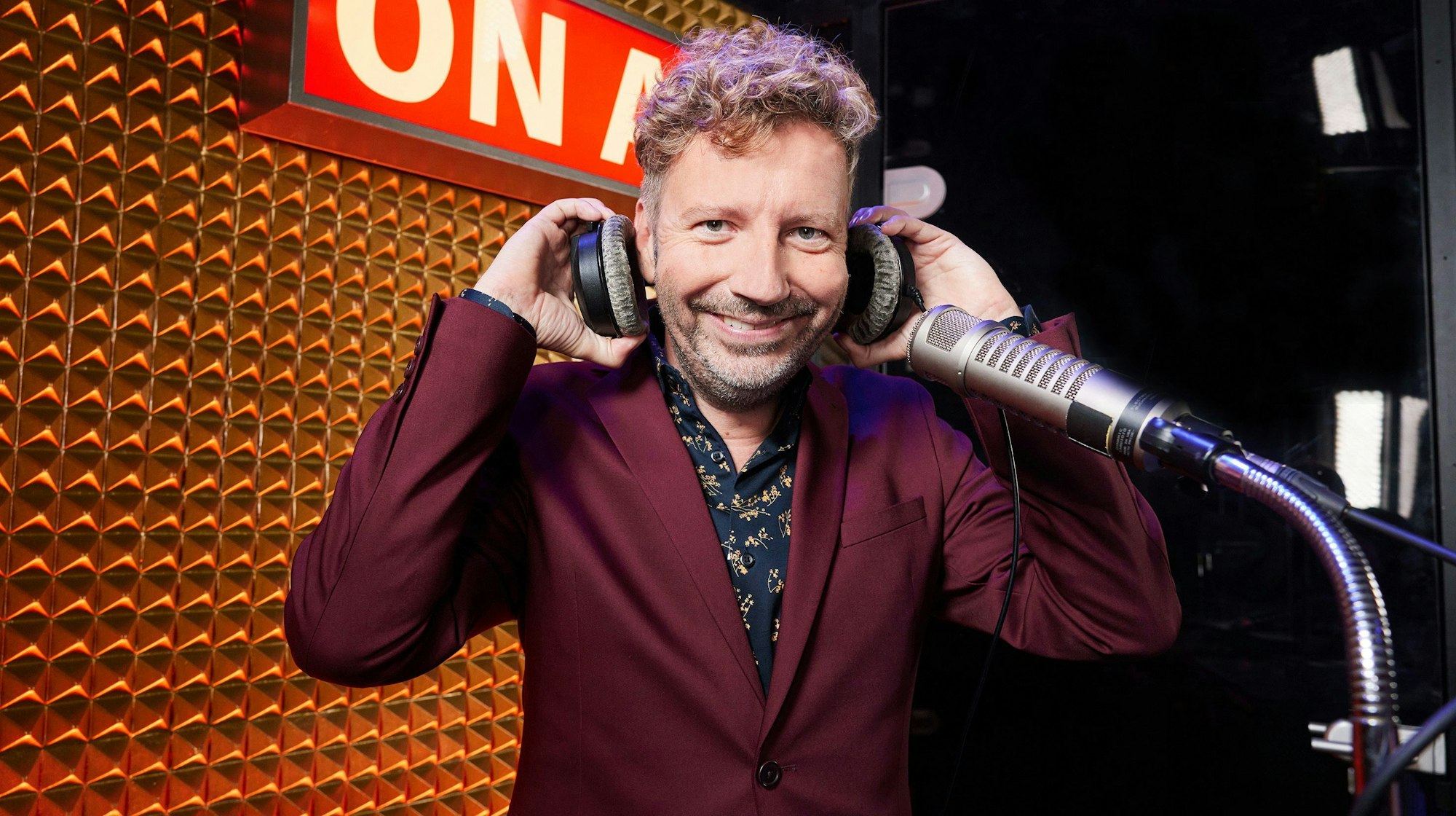 Thorsten Schorn steht an einem Mikrofon, er hat Kopfhörer in der Hand und trägt einen bordeauxfarbenen Anzug.
