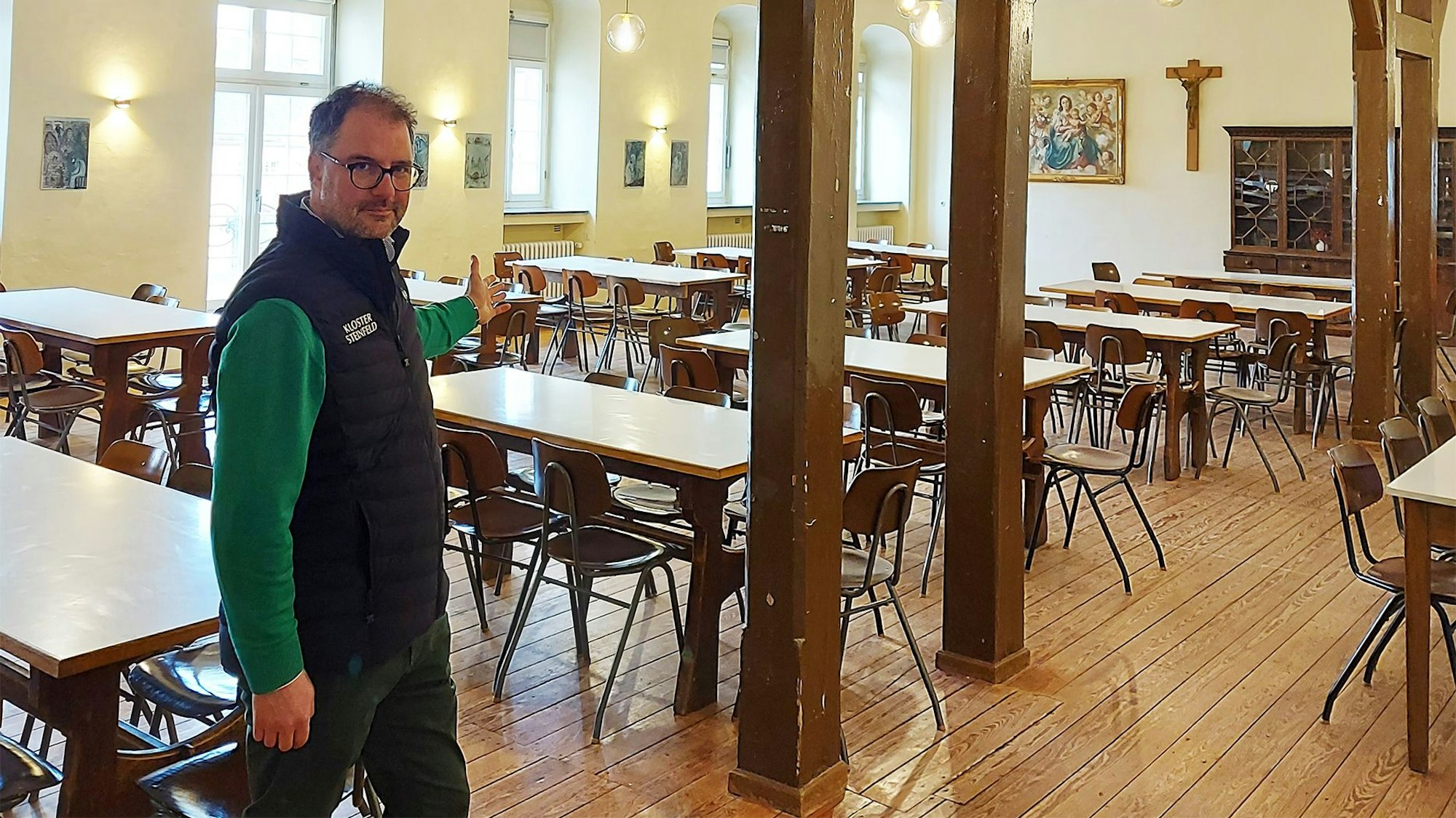 Christoph Böhnke zeigt in den Hermann-Josef-Saal im Kloster Steinfeld, in dem zahlreiche Tische und Stühle stehen.