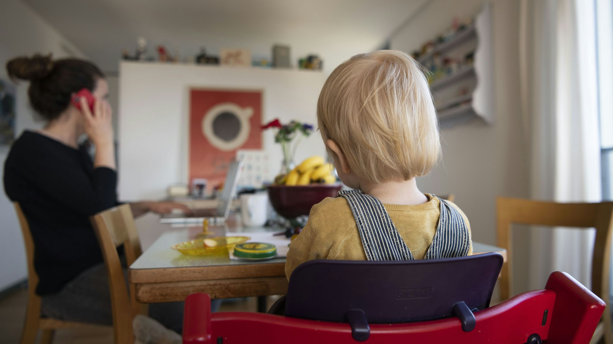 Eine Frau sitzt im Homeoffice an ihrem Laptop und telefoniert, während ihr Kind neben ihr in einem Kinderstuhl am Tisch sitzt.