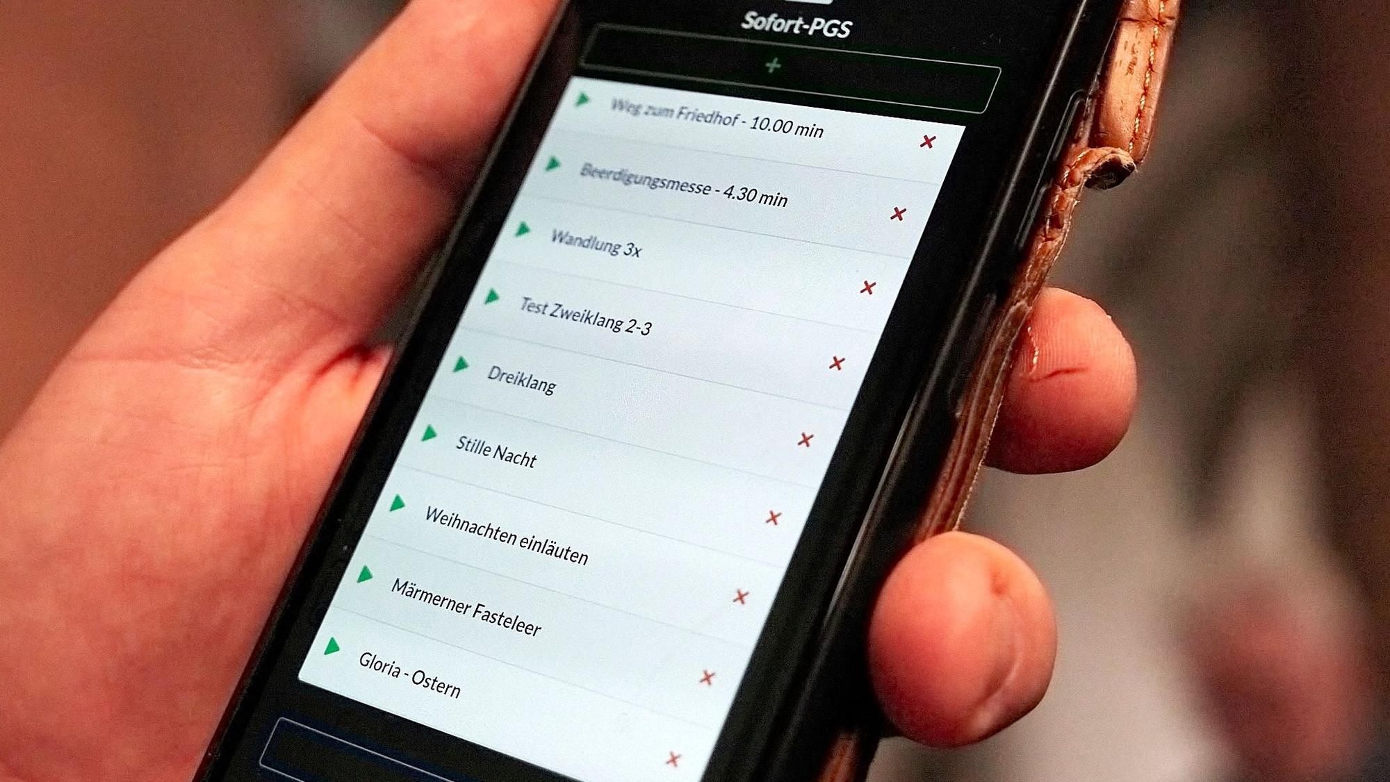 In einer Hand wird ein Smartphone gehalten, auf dessen Bildschirm verschiedene Optionen zu sehen sind. Sie können gewählt werden, um die Glocken der Kirche in Marmagen zu steuern.