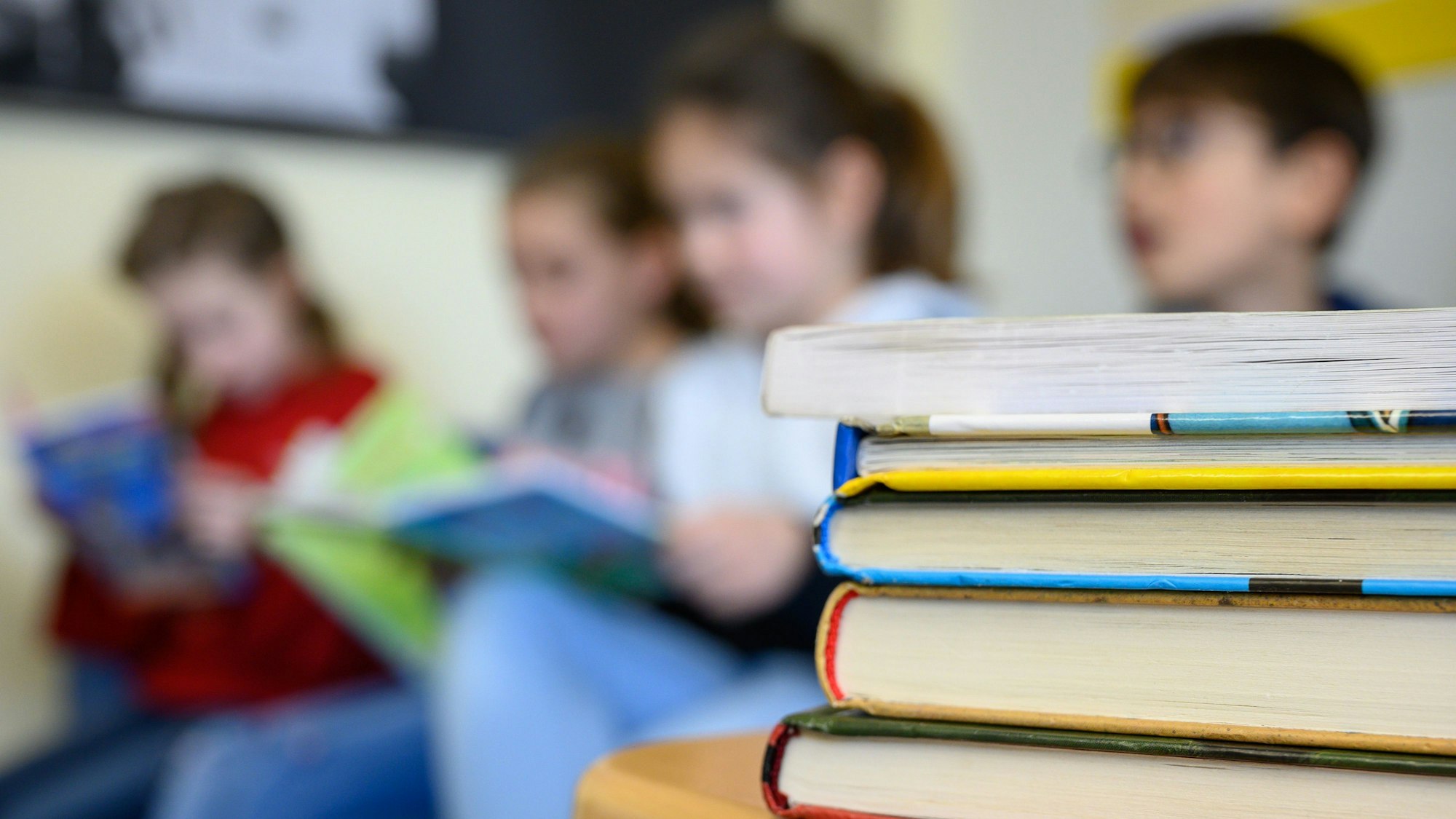 Im Vordergrund liegt ein Stapel Bücher, im Hintergrund lesen Kinder in einer Grundschule.