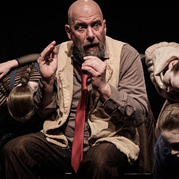 Das Bild zeigt den Schauspieler Thomas Balou Martin. Er trägt eine Glatze und einen Bart. Er hat eine Weste an, darunter ein braunes Hemd und eine lange, rote Krawatte. Weitere Schauspielerinnen tummeln sich um ihn herum.