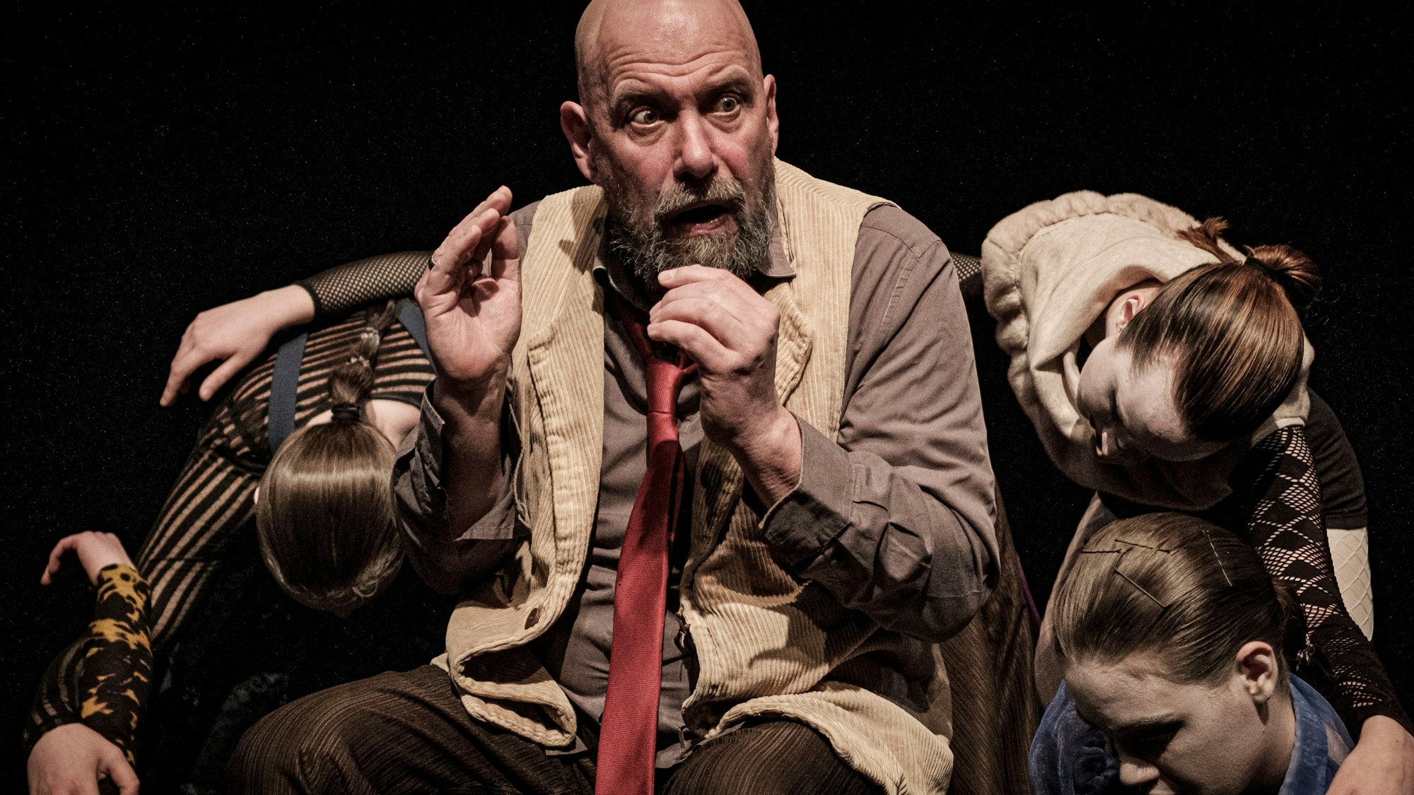 Das Bild zeigt den Schauspieler Thomas Balou Martin. Er trägt eine Glatze und einen Bart. Er hat eine Weste an, darunter ein braunes Hemd und eine lange, rote Krawatte. Weitere Schauspielerinnen tummeln sich um ihn herum.