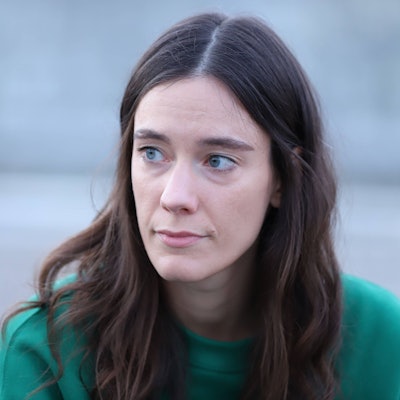 Auf dem Bild ist die TikTokerin ‚Toxische Pommes‘ bis auf Höhe der Schultern vor einem grauen, unscharf gemachten Hintergrund zu sehen. ‚Toxische Pommes‘ blickt nicht in die Kamera, sondern nach links.