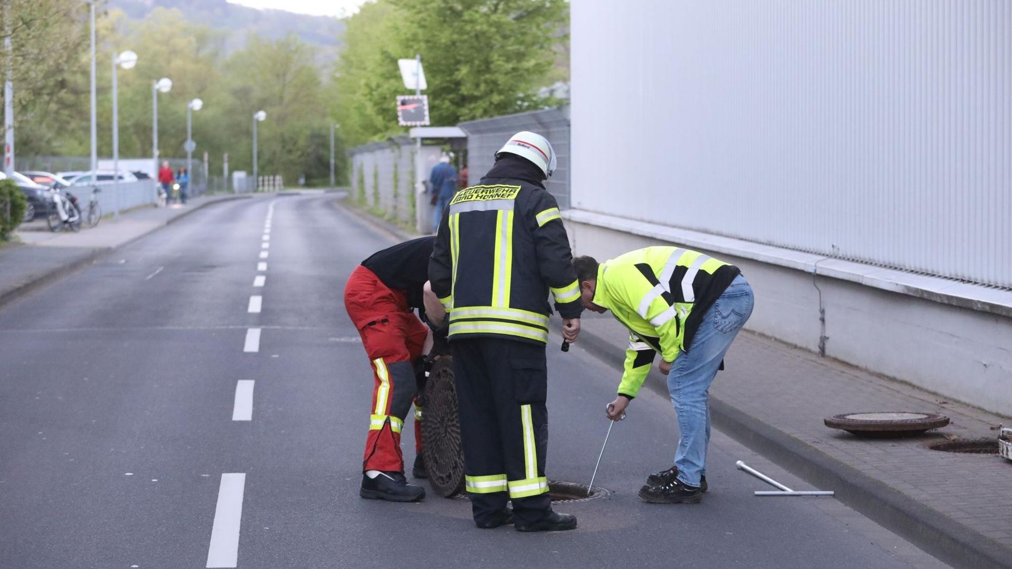 Drei Männer, darunter ein Feuerwehrmann, stehen auf einer Straße und schauen in den geöffneten Kanalschacht.