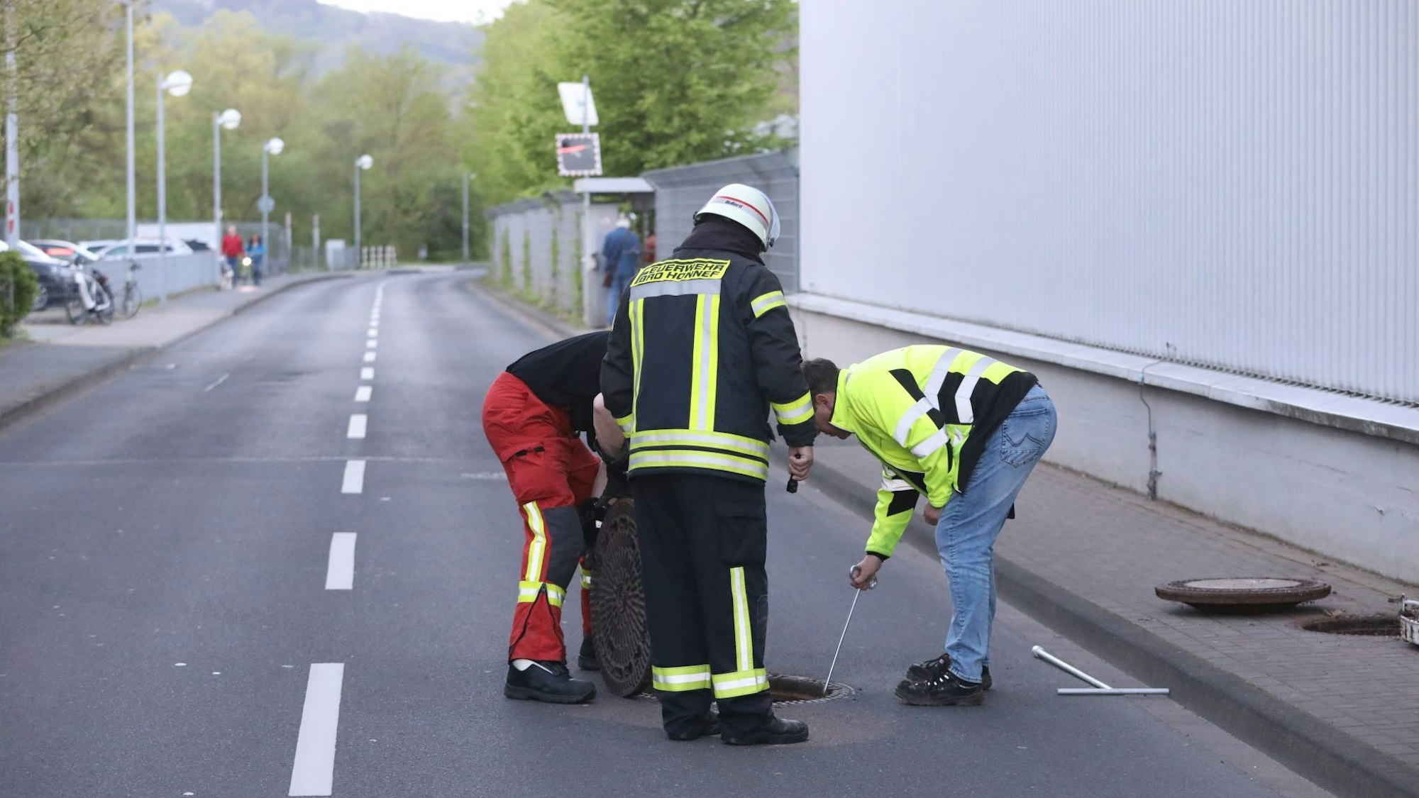 Drei Männer, darunter ein Feuerwehrmann, stehen auf einer Straße und schauen in den geöffneten Kanalschacht.