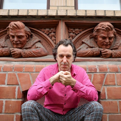 Chilly Gonzales trägt ein pinkes Hemd und hockt auf der Richard-Wagner-Straße vor zwei Reliefbüsten.