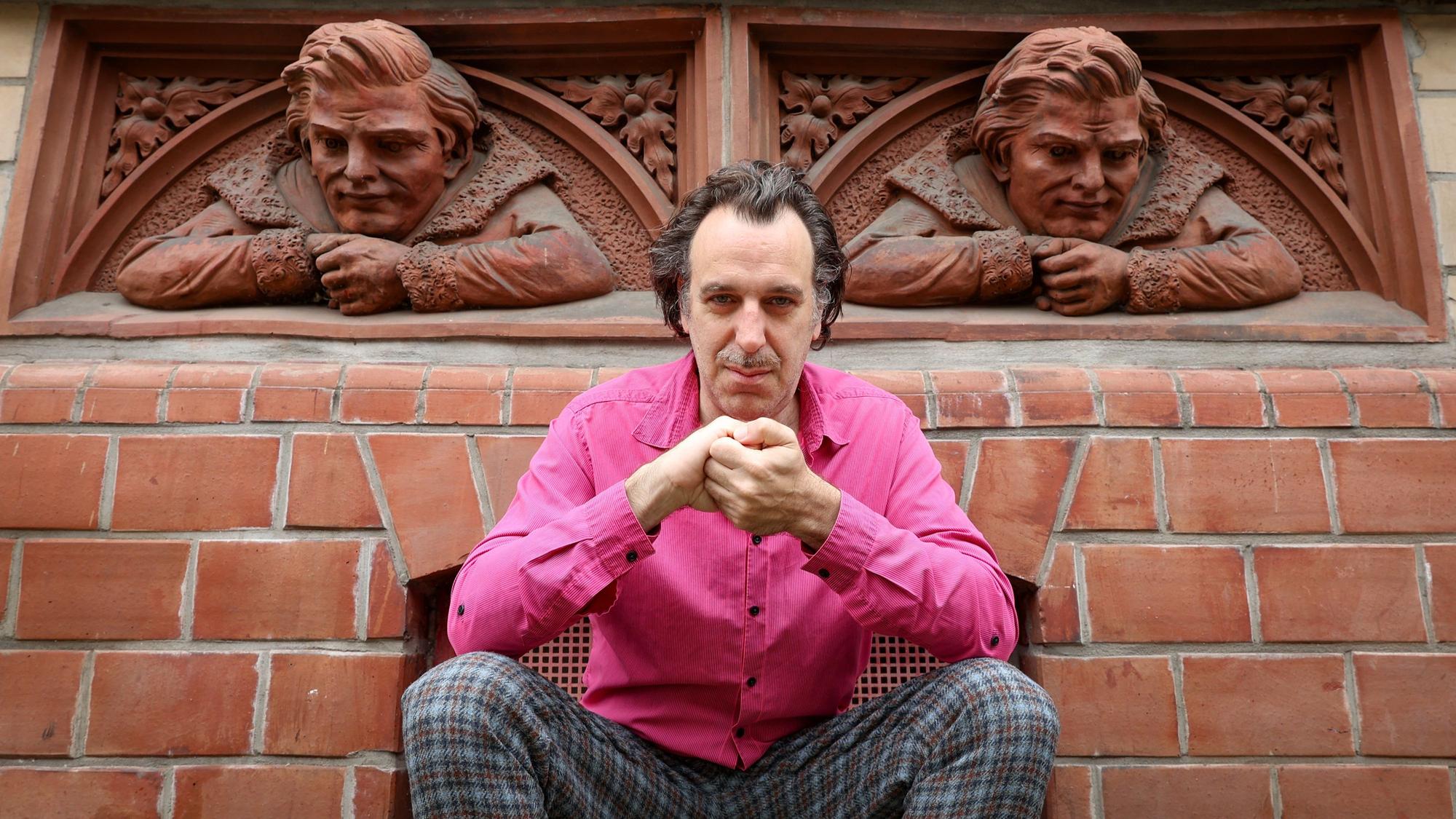 Chilly Gonzales trägt ein pinkes Hemd und hockt auf der Richard-Wagner-Straße vor zwei Reliefbüsten.