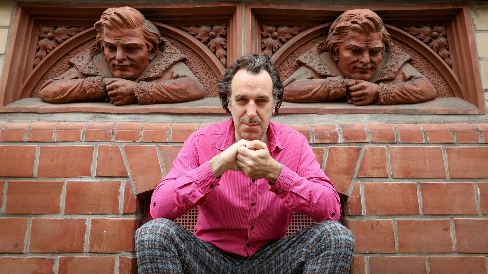 Chilly Gonzales trägt ein pinkes Hemd und hockt auf der Richard-Wagner-Straße vor zwei Reliefbüsten.