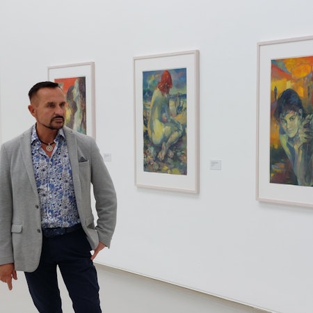 Ein Mann im grauen Sakko und einem weiß-blau gemusterten Hemd in einem Museumssaal; an der Wand hängen farbenfrohe Bilder.