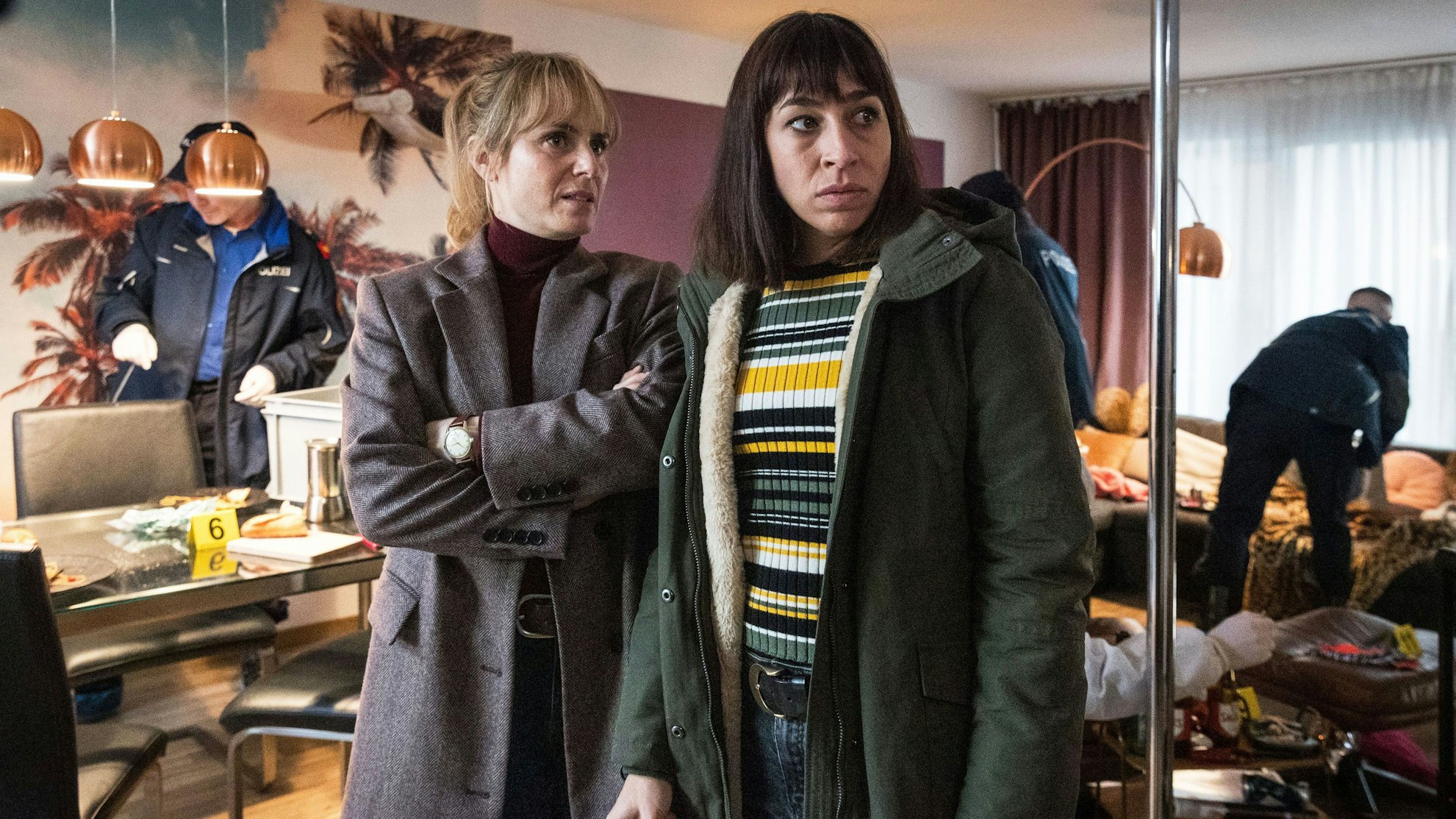 Im Vordergrund stehen die beiden Kommissarinnen Isabelle Grandjean und Tessa Ott und schauen skeptisch in Richtung Tatort. Im Hintergrund sieht man, wie Polizeikollegen die Wohnung gründlich unter die Lupe nehmen und nach Spuren untersuchen.