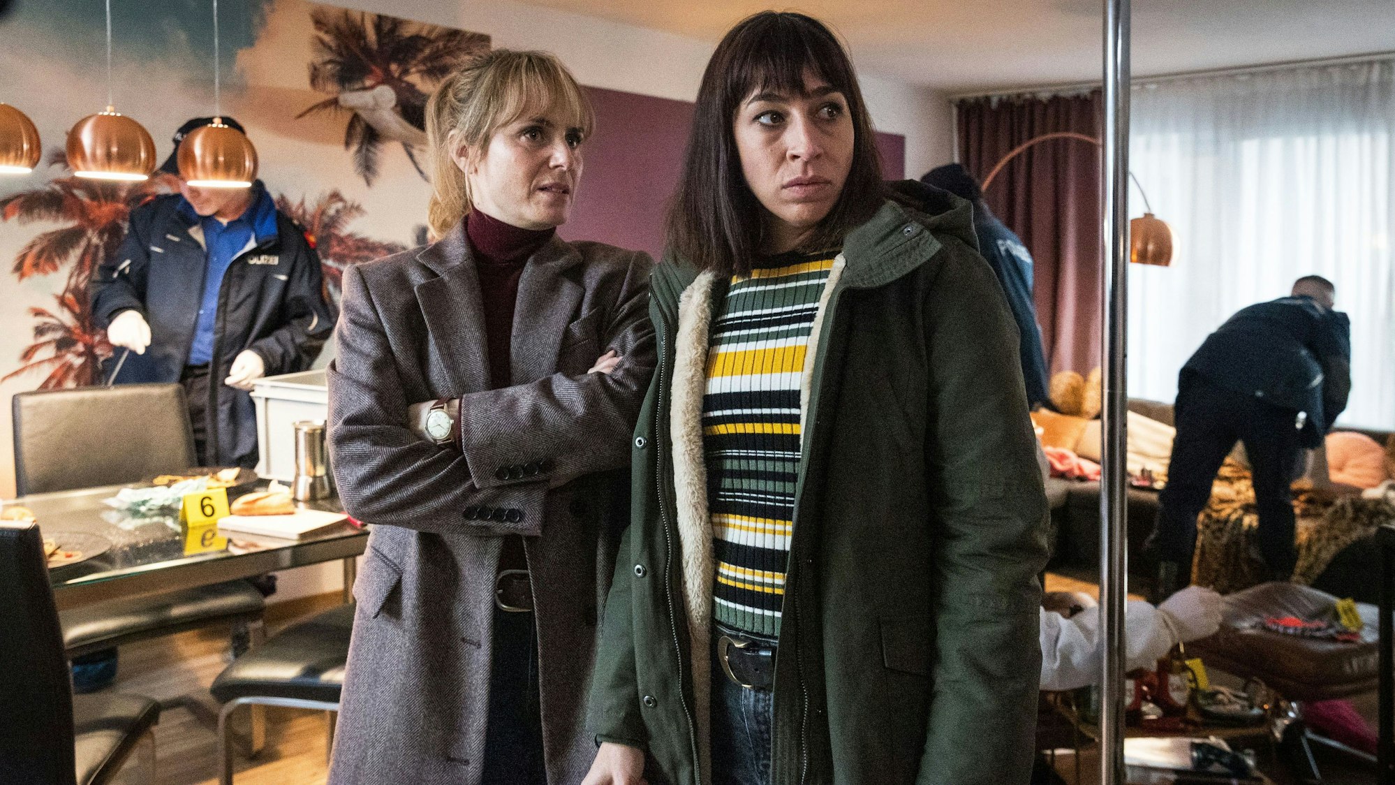 Im Vordergrund stehen die beiden Kommissarinnen Isabelle Grandjean und Tessa Ott und schauen skeptisch in Richtung Tatort. Im Hintergrund sieht man, wie Polizeikollegen die Wohnung gründlich unter die Lupe nehmen und nach Spuren untersuchen.