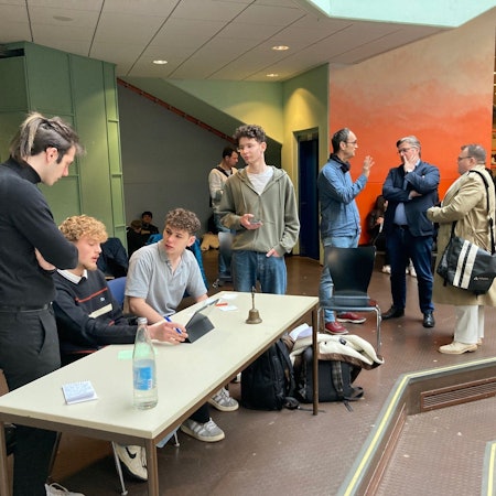 An einem Tisch sitzen die drei Moderatoren und bereiten sich vor. Im Hintergrund stehen Lehrer und zwei Kandidaten und unterhalten sich.