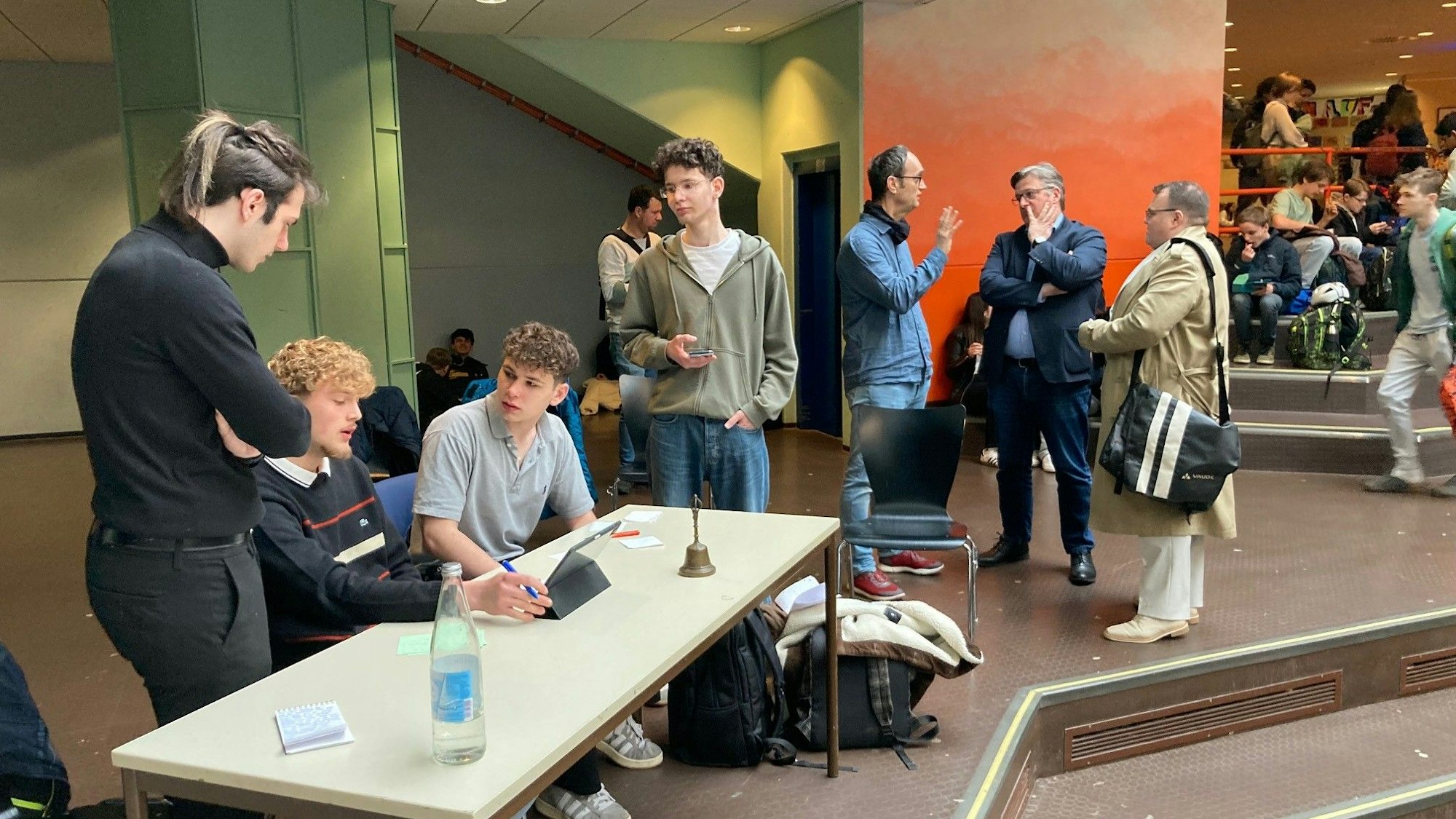 An einem Tisch sitzen die drei Moderatoren und bereiten sich vor. Im Hintergrund stehen Lehrer und zwei Kandidaten und unterhalten sich.