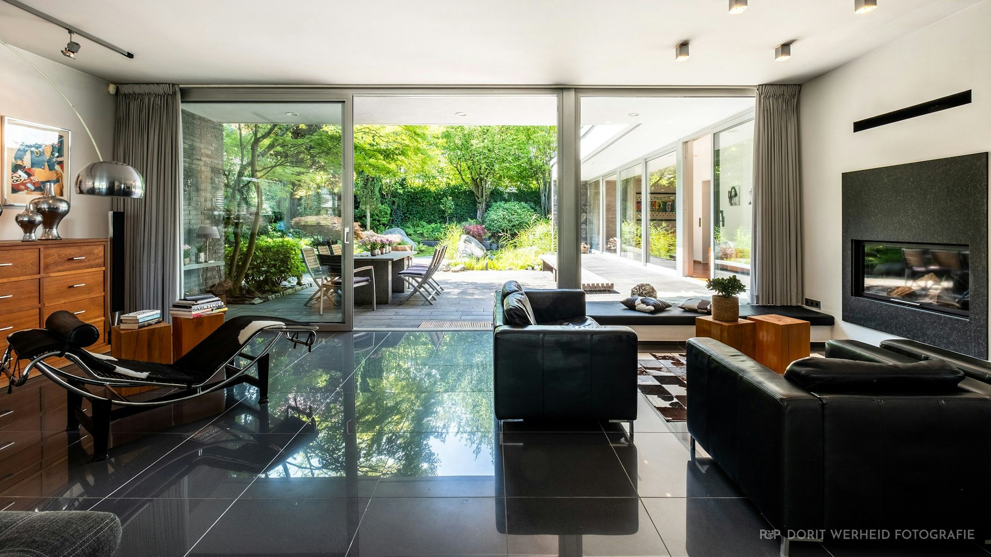 Das Wohnzimmer der Immobilie in Königsdorf, ausgestattet unter anderem mit dunklen Möbeln. Im Hintergrund ist der Zugang zu einer der Terrassen und den angrenzenden Garten zu sehen.