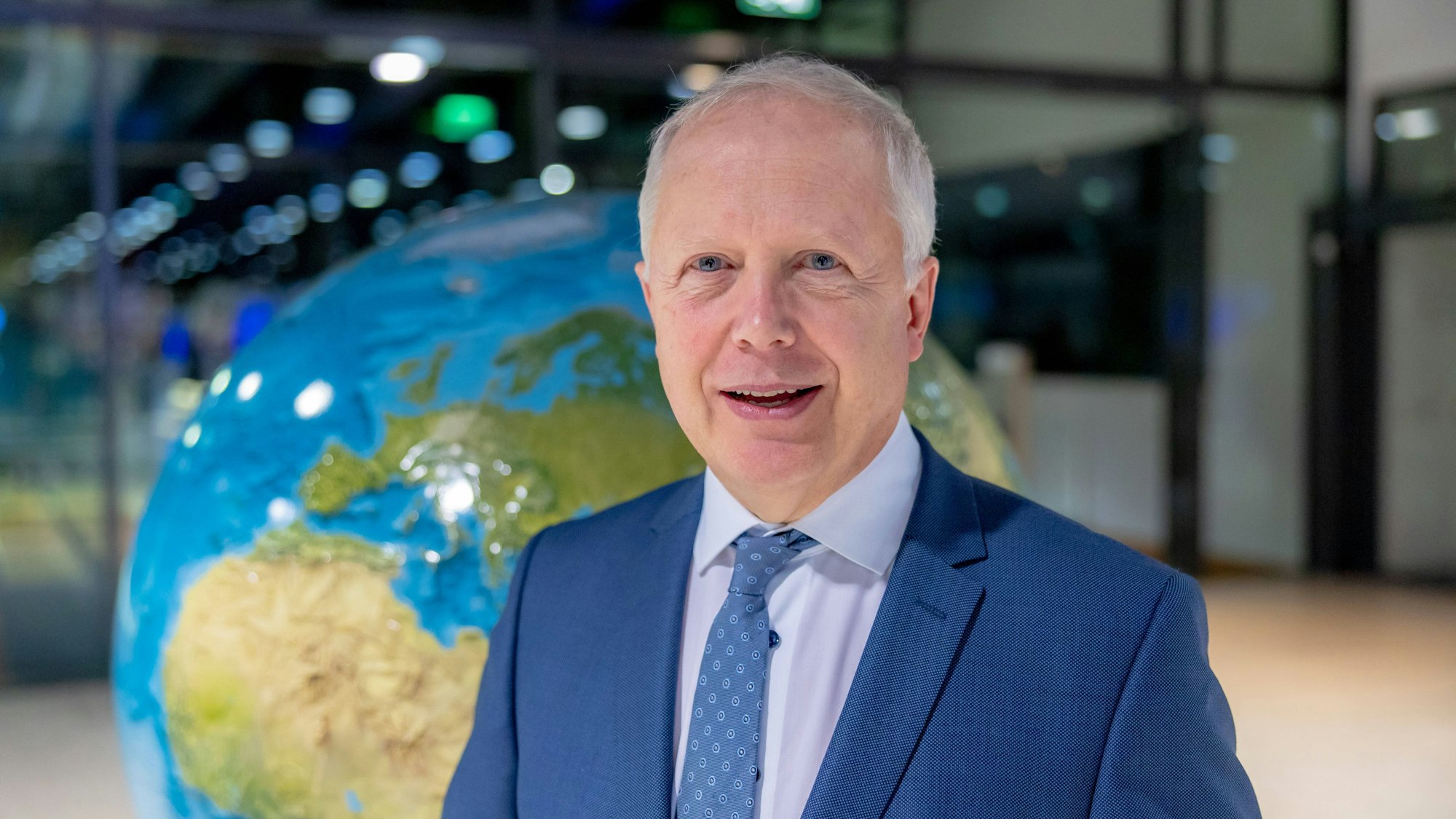 WDR-Intendant Tom Buhrow steht vor einer großen Weltkugel. Er trägt einen blauen Anzug und eine blaue Krawatte.
