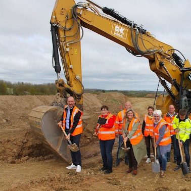 Vor einem Bagger wird der erste Spatenstich vollzogen.