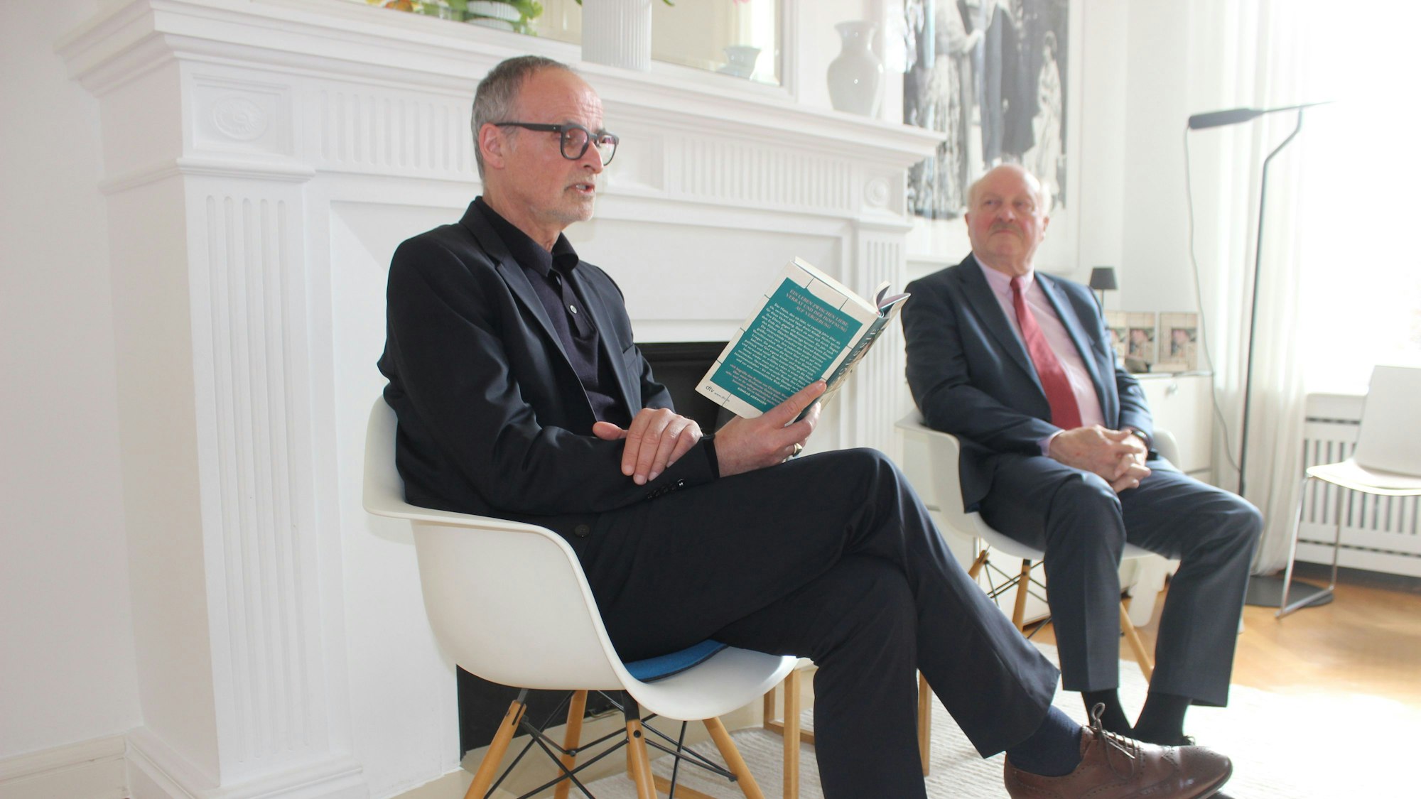 Zwei Männer sitzen auf Stühlen, einer liest aus einem Buch.