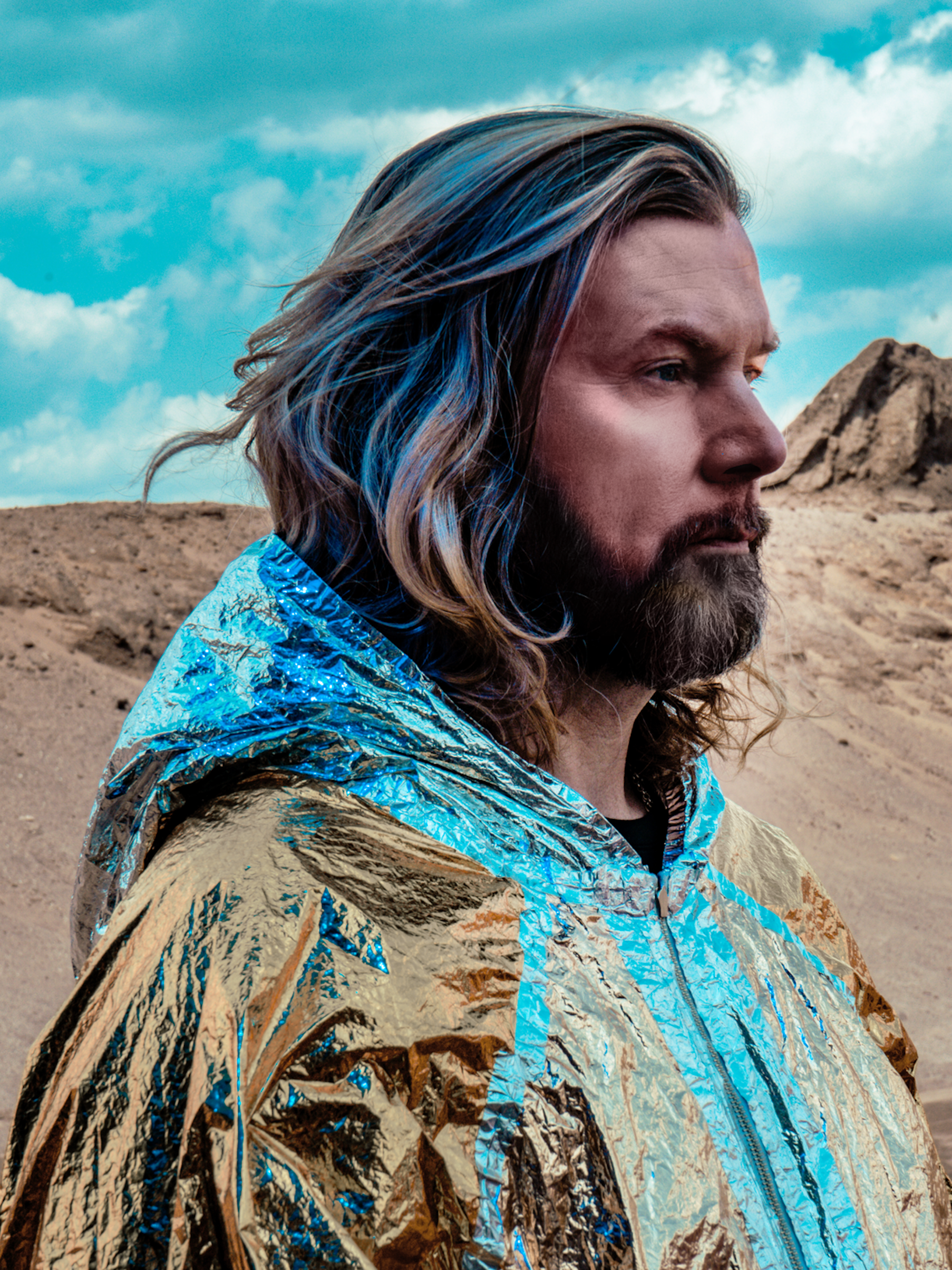 Porträt von Rea Garvey von der Seite. Er trägt einen gold-hellblauen Umhang, die Haar sind teilweise mit blauen Strähnen durchsetzt