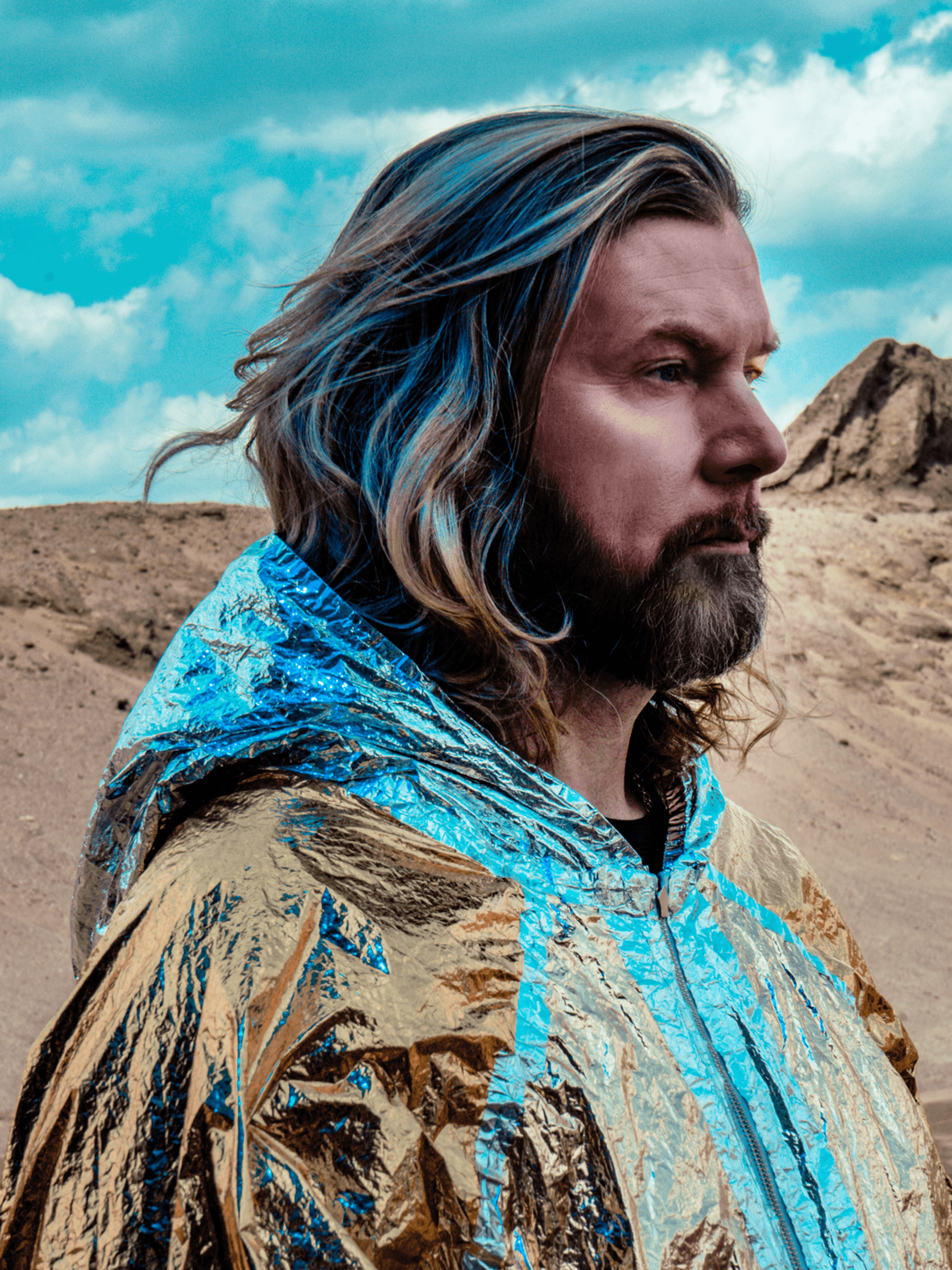 Porträt von Rea Garvey von der Seite. Er trägt einen gold-hellblauen Umhang, die Haar sind teilweise mit blauen Strähnen durchsetzt