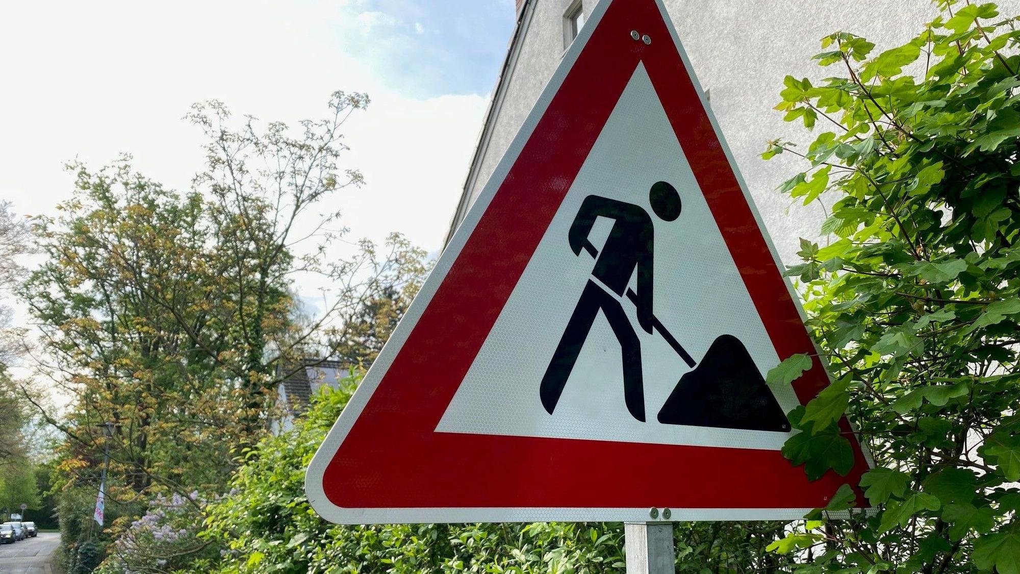 Ein Baustellenschild steht an einer Hecke. Im Hintergrund ist eine Straße zu sehen..