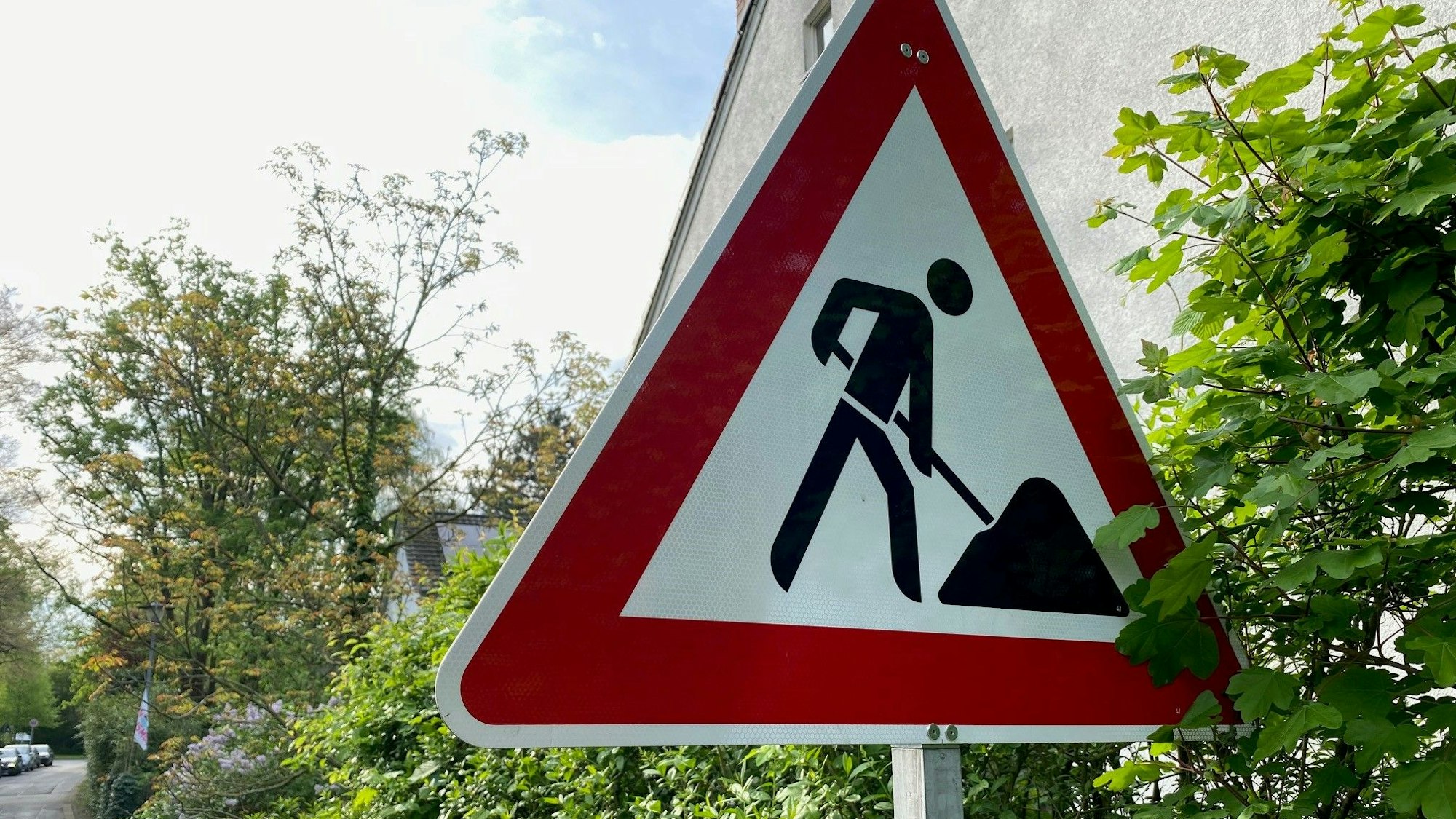 Ein Baustellenschild steht an einer Hecke. Im Hintergrund ist eine Straße zu sehen..