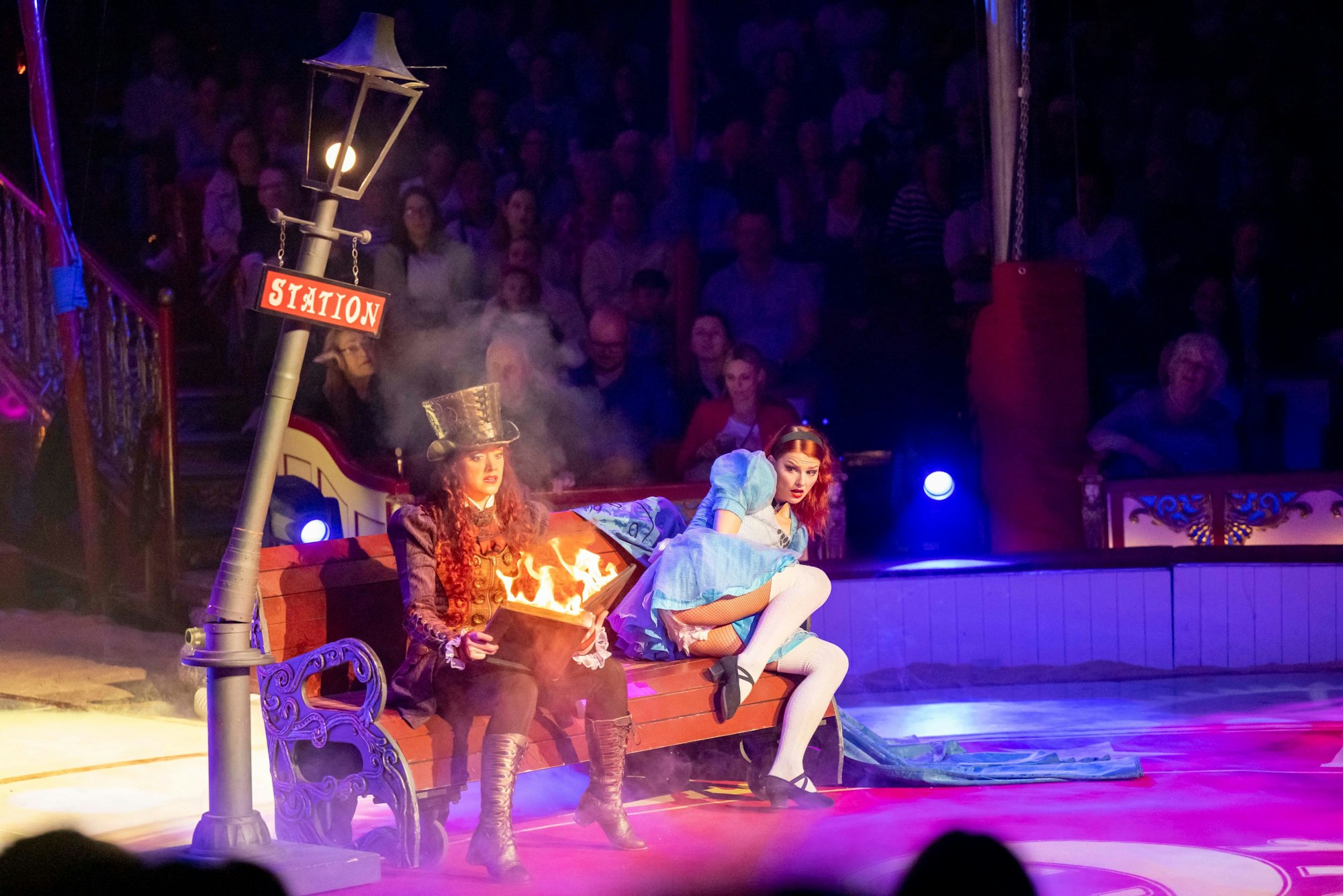 Zwei Frauen auf einer Bank, eine hält ein brennendes Buch, die andere ist als Alice im Wunderland verkleidet.