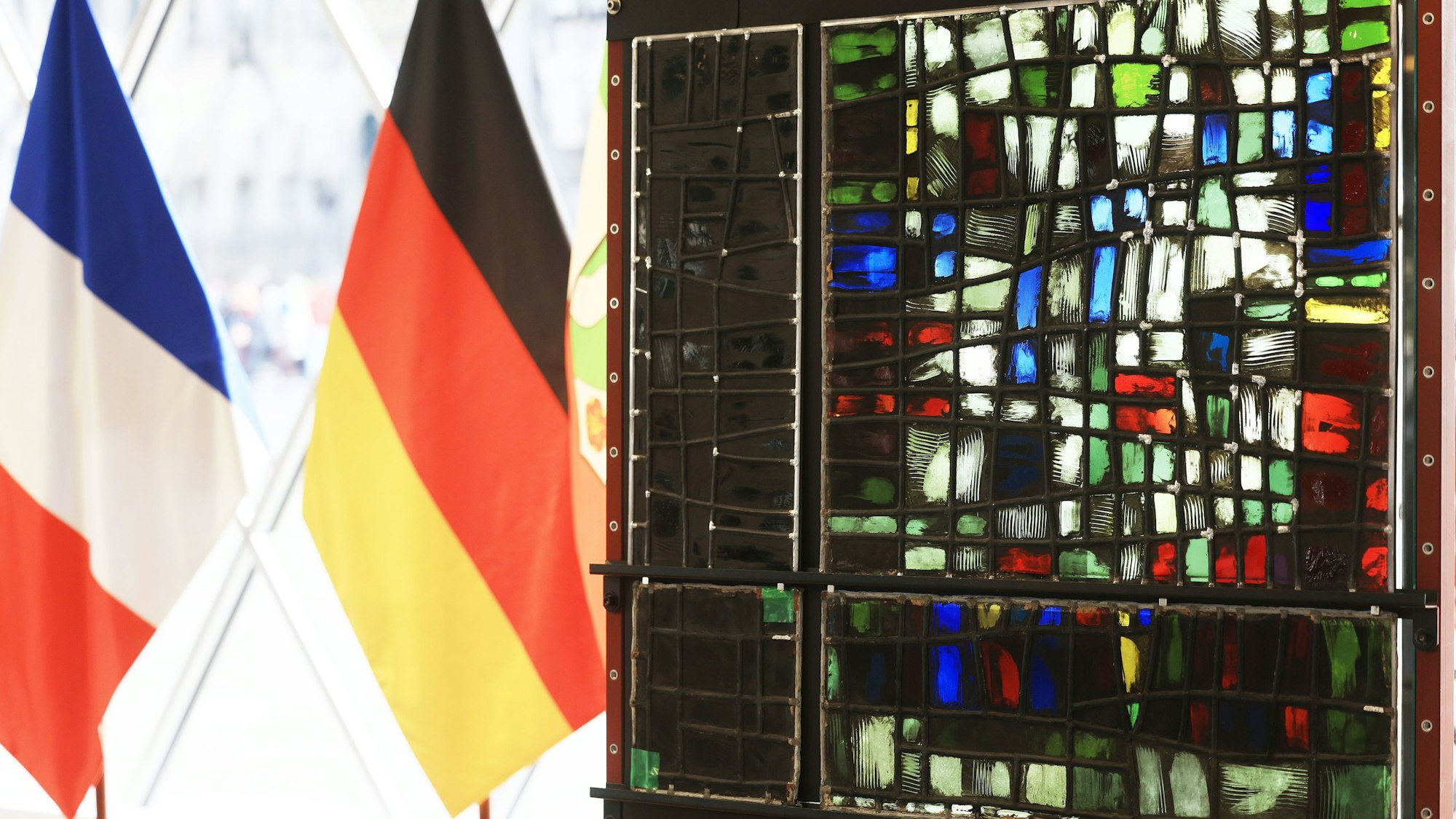 Der restaurierte (r.) und verrußte Teil (l.) eines Fenster aus der Kirche Notre-Dame hängen neben der deutschen und französischen Fahne im Kölner Domforum. Die Wiederherstellung der Glaskunst wurde als Symbol deutsch-französischer Zusammenarbeit verstanden.