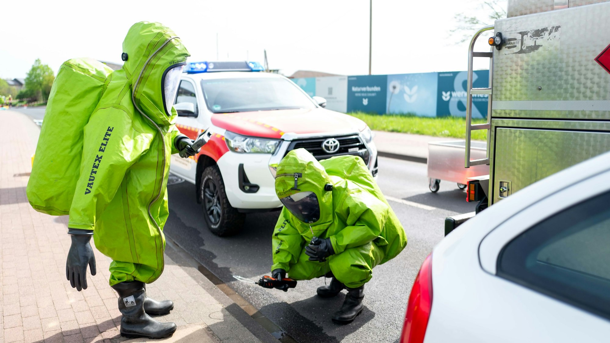 Das Bild zeigt zwei Feuerwehrleute, die einen grünen Schutzanzug anhaben.