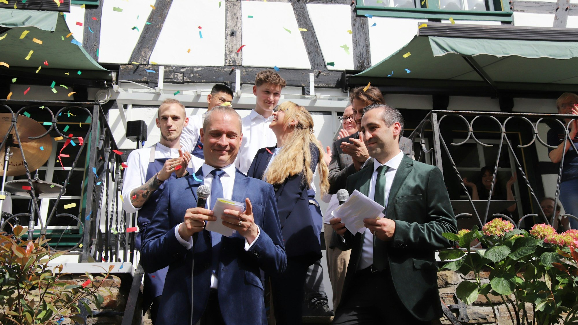 Zwei Männer stehen im Konfettiregen vor einem Fachwerkhaus, Servicepersonal ist hinter ihnen zu sehen.